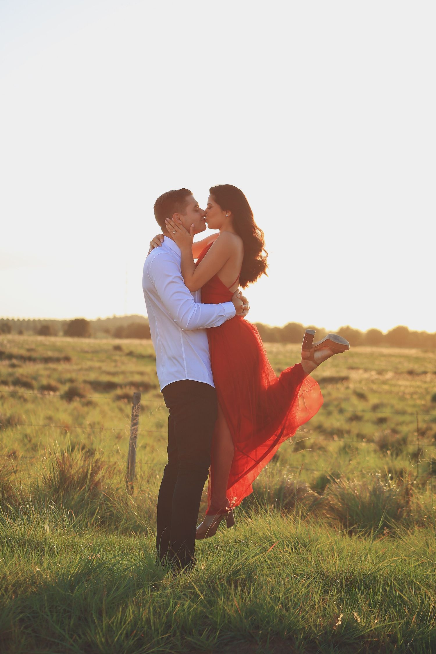 Romantic sunset photo of couple embracing in a field with flowing red dress catching the golden light.