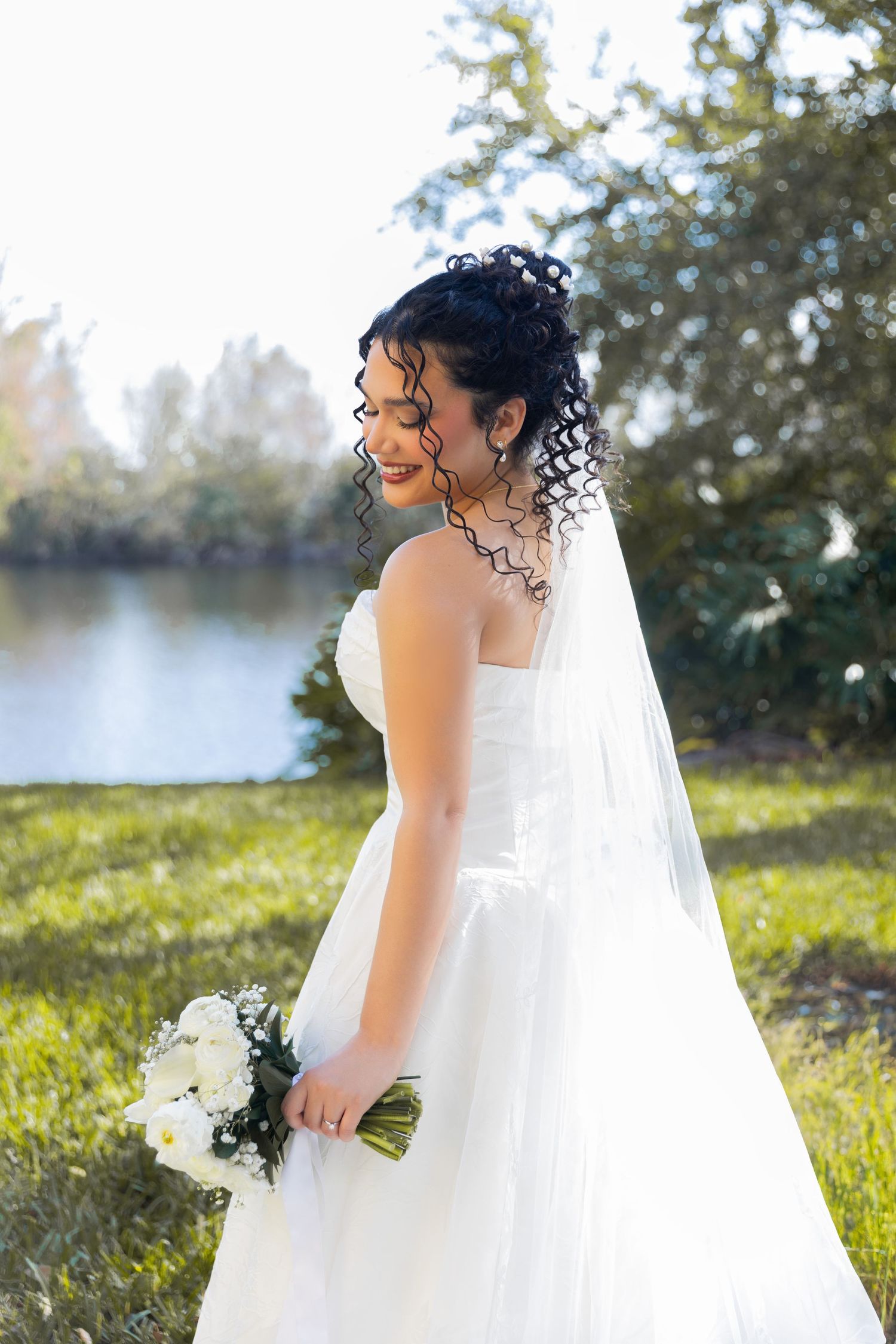 A bride in a flowing white wedding dress stands in a sunlit outdoor setting near a lake.
