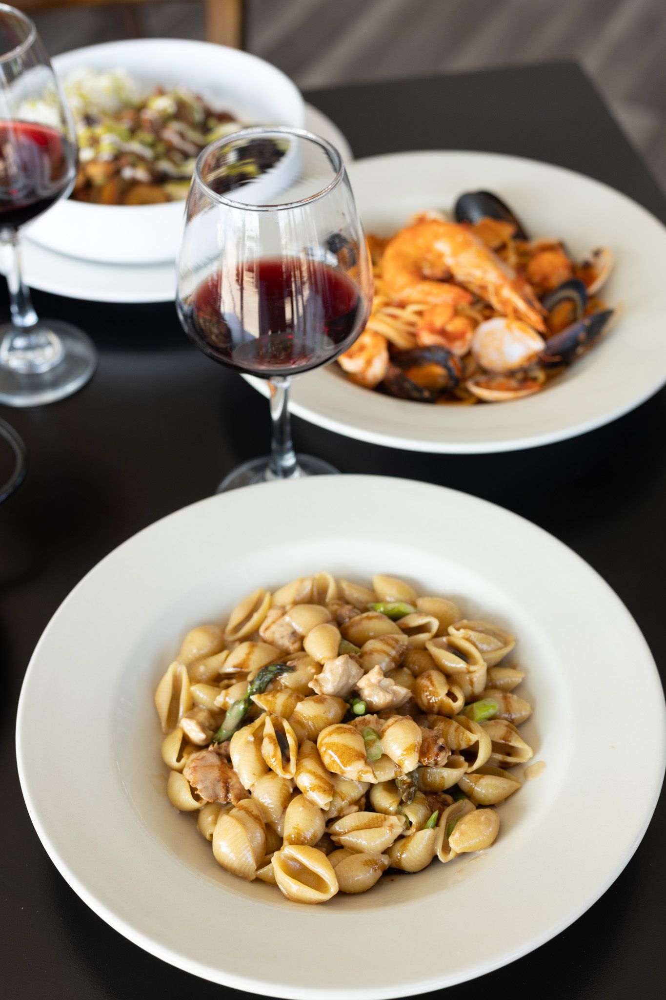 Elegant white plates of orecchiette pasta with seafood and wine glasses on a dark table setting.