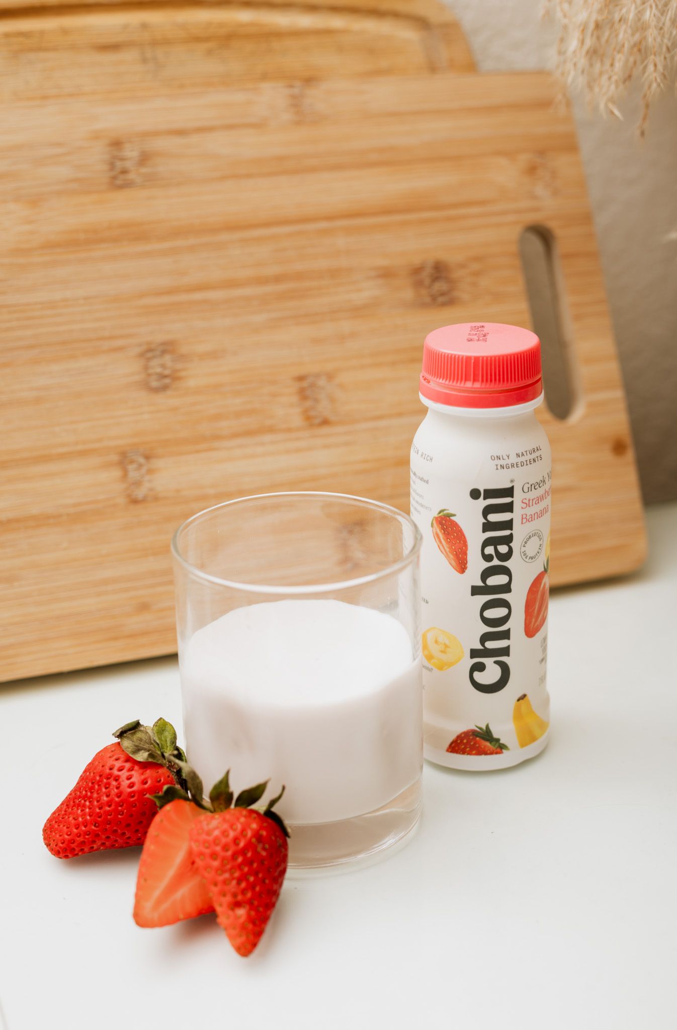 Chobani strawberry banana yogurt drink beside a glass of yogurt and fresh strawberries on a white surface.