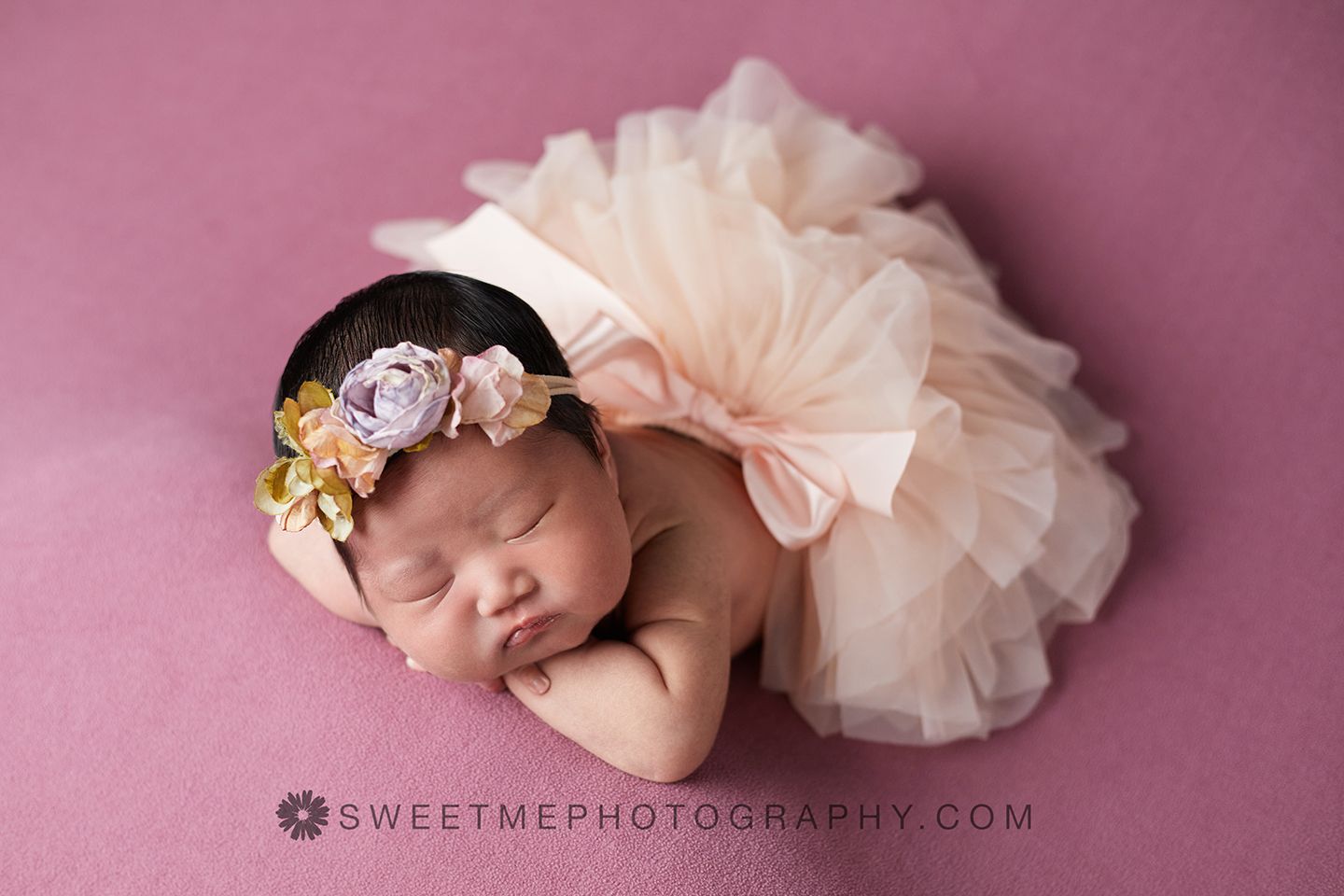 A sleeping newborn baby wearing a pink tulle dress and floral headband rests on a pink backdrop during a professional photoshoot.