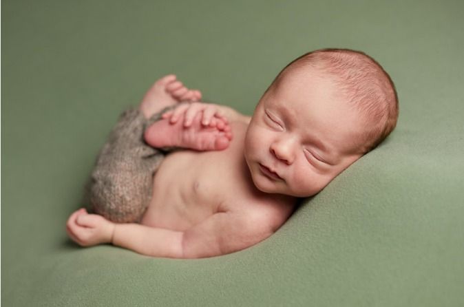 Sleeping newborn baby curled up peacefully on soft sage green backdrop wearing knit pants.
