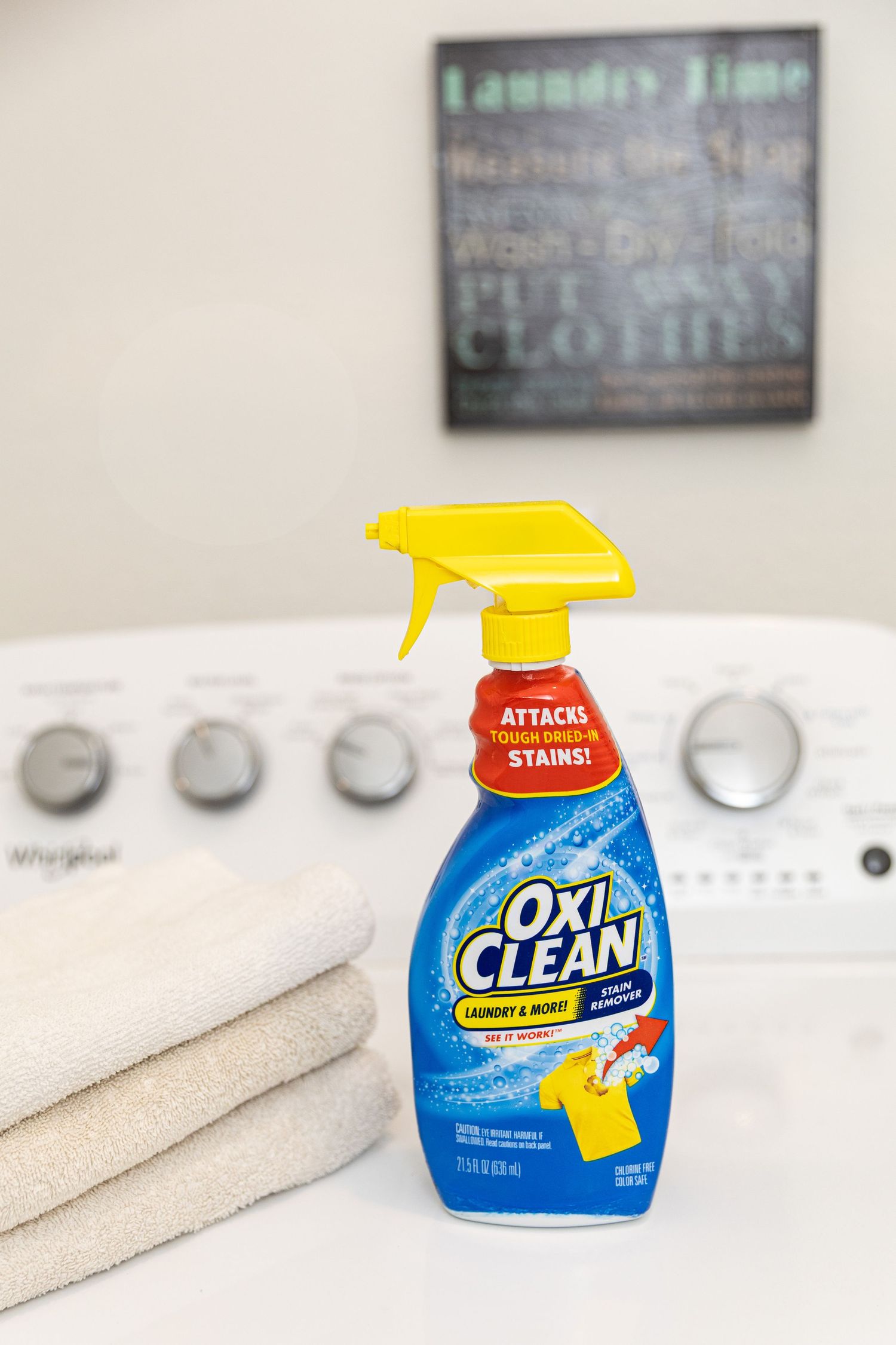 OxiClean laundry stain remover spray bottle on a washing machine with folded towels in a laundry room.