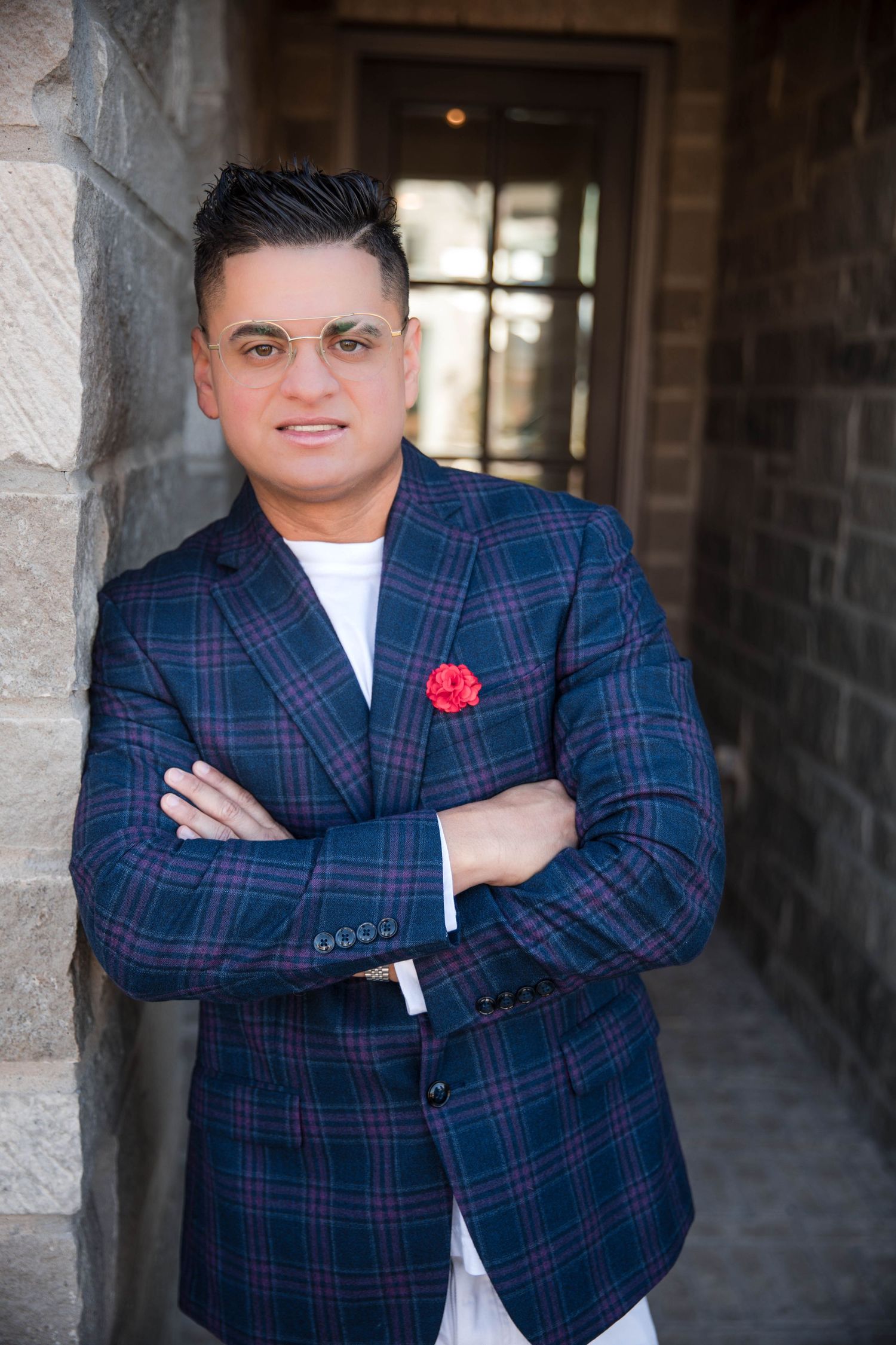 A person wearing a navy plaid blazer with a red boutonniere poses in a stone archway.