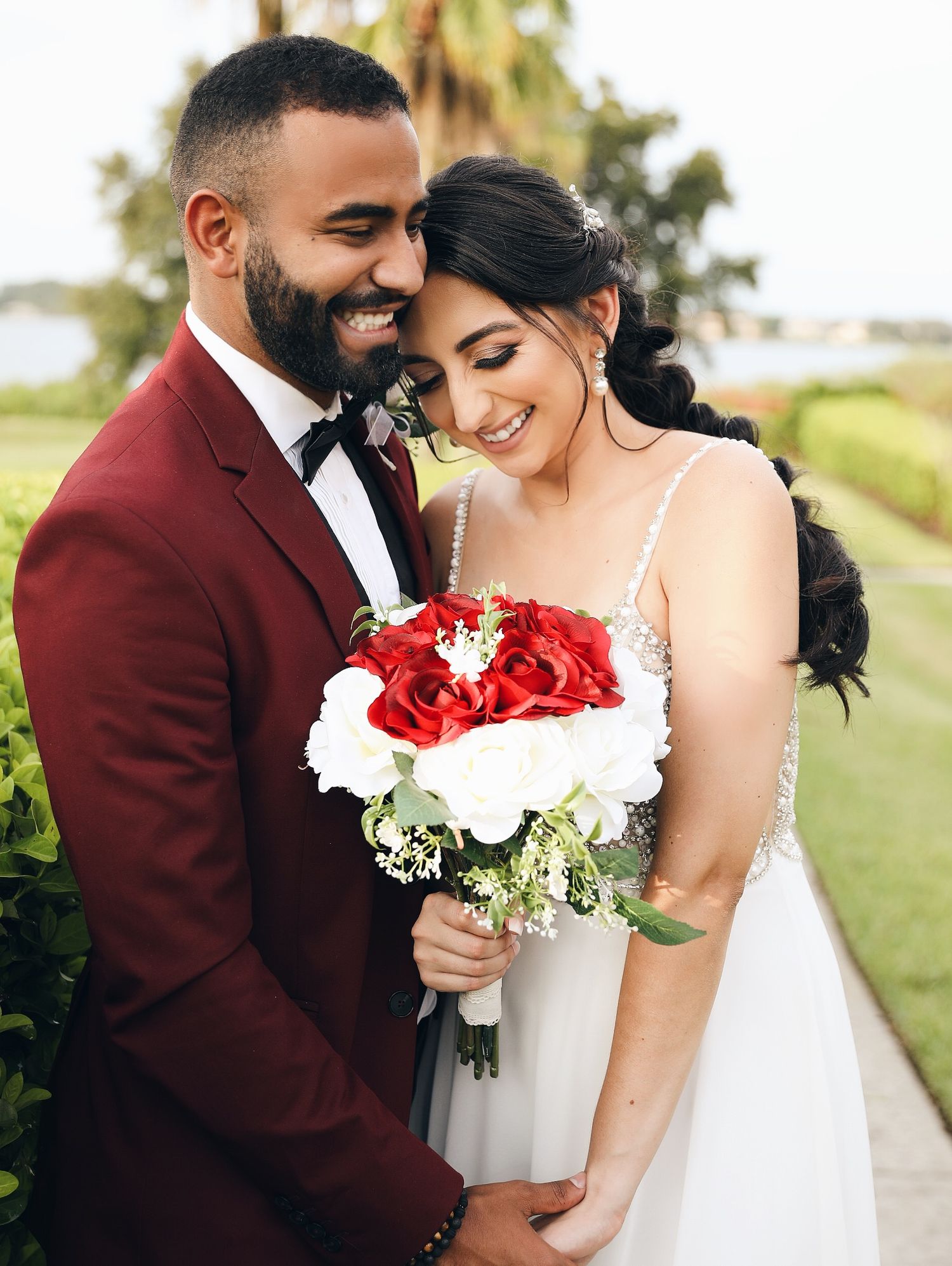 A romantic wedding couple shares intimate moments in a garden setting wearing burgundy and white attire with red bouquet.
