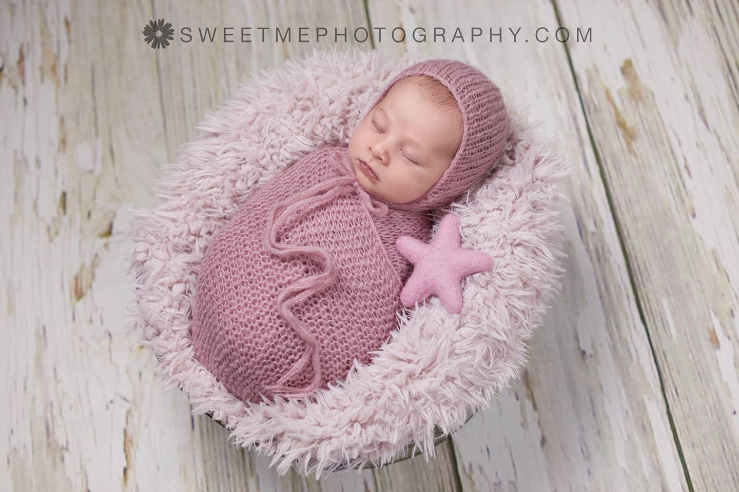 A sleeping newborn in pink swaddle and knit wrap lies on a textured white wooden backdrop during a photography session.