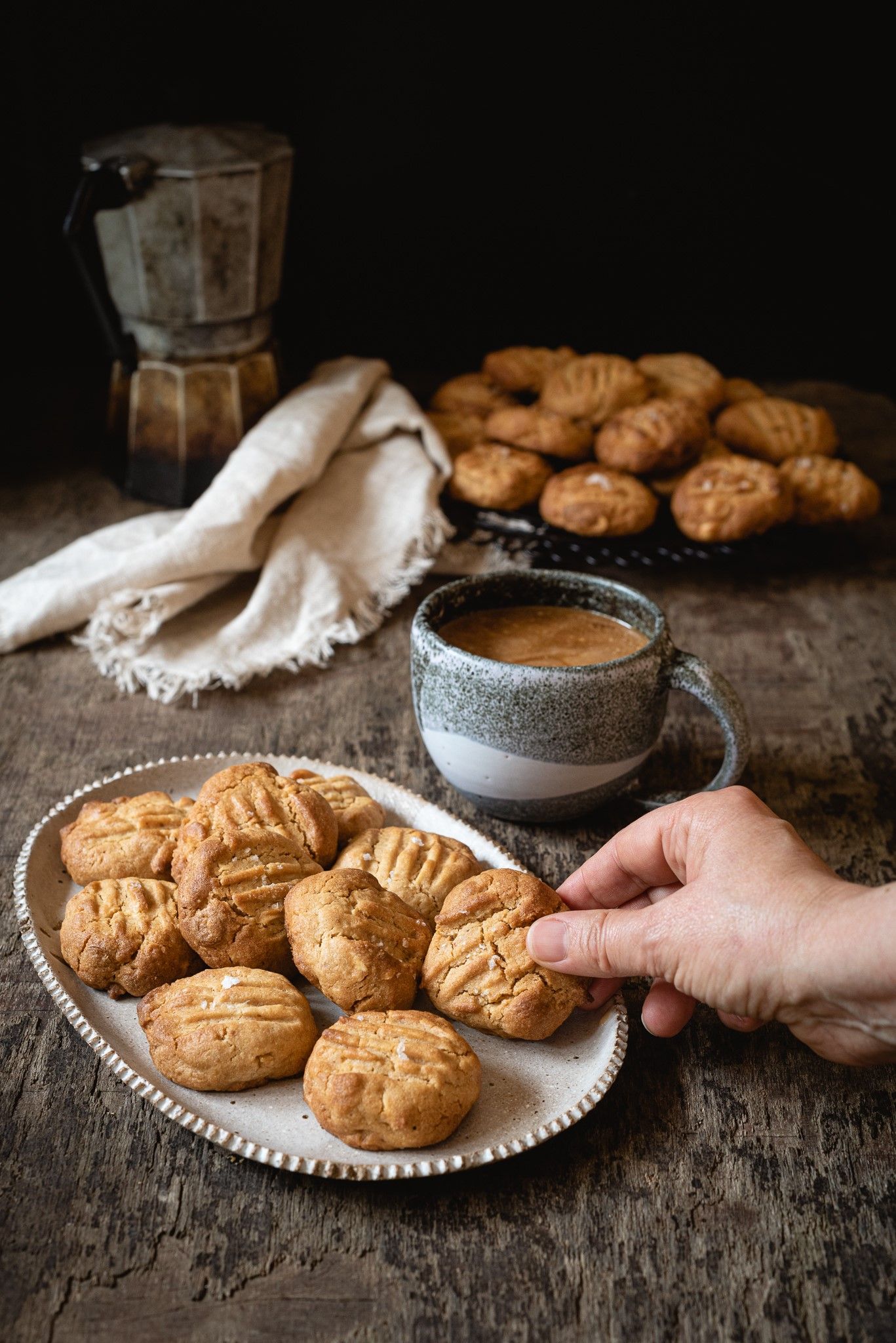 Hand reaching for peanut butter cookies on a plate, with coffee cup and moka pot on rustic wooden table.