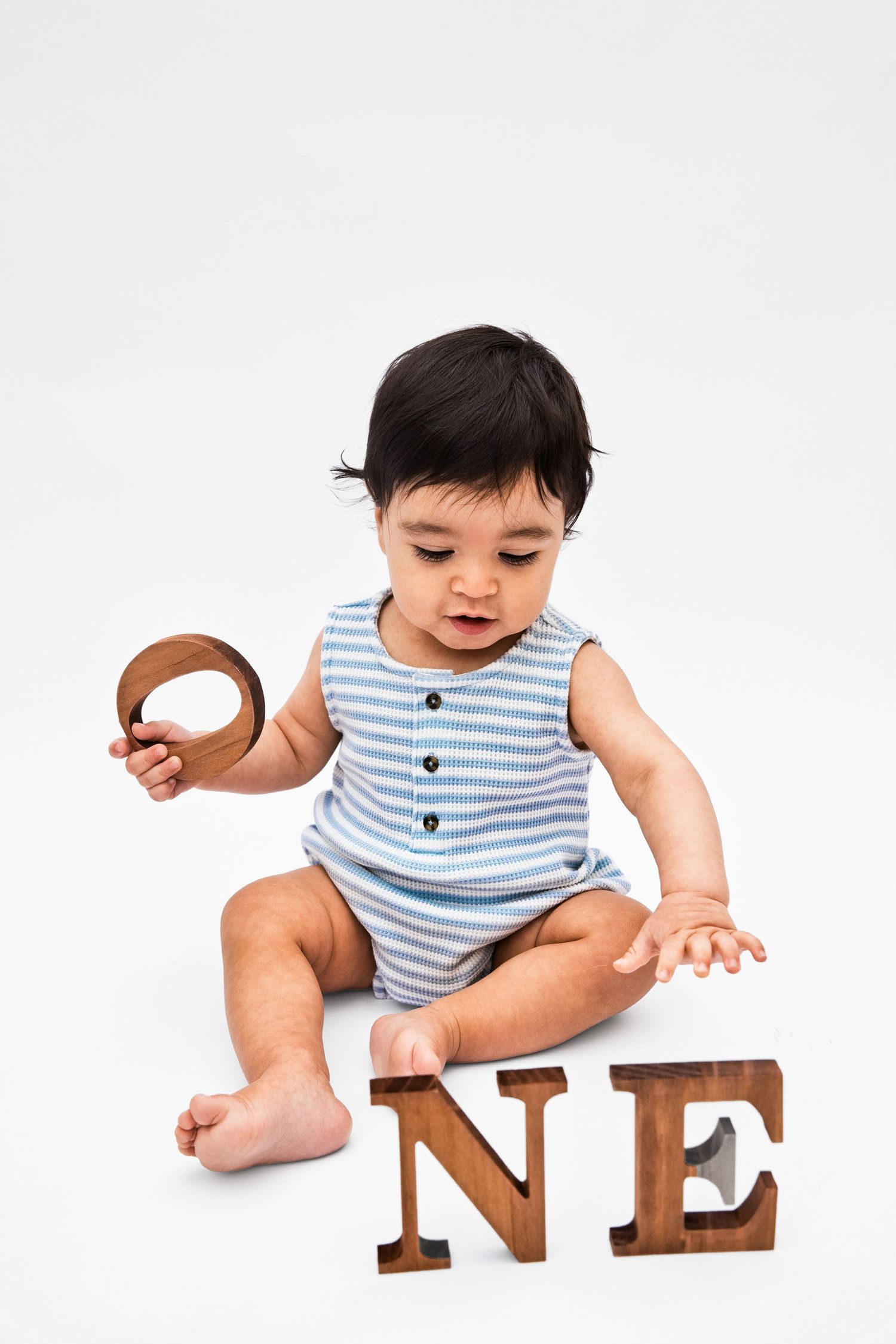 Cute baby in striped romper sitting with wooden letters spelling ONE, celebrating first birthday on white background.