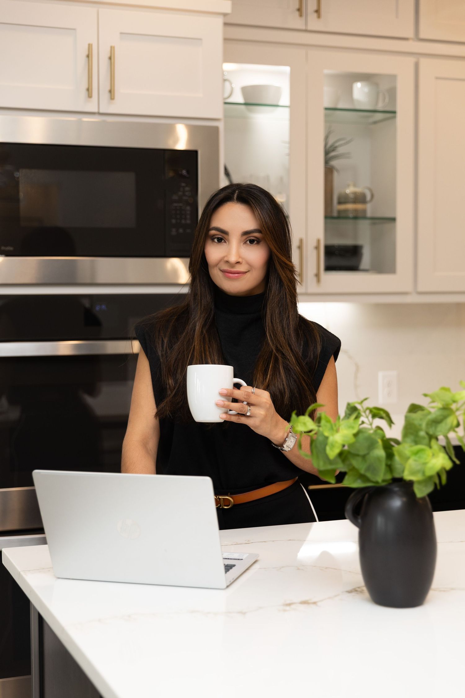 Woman in black outfit holding coffee cup at kitchen island with laptop and plant in modern white kitchen.
