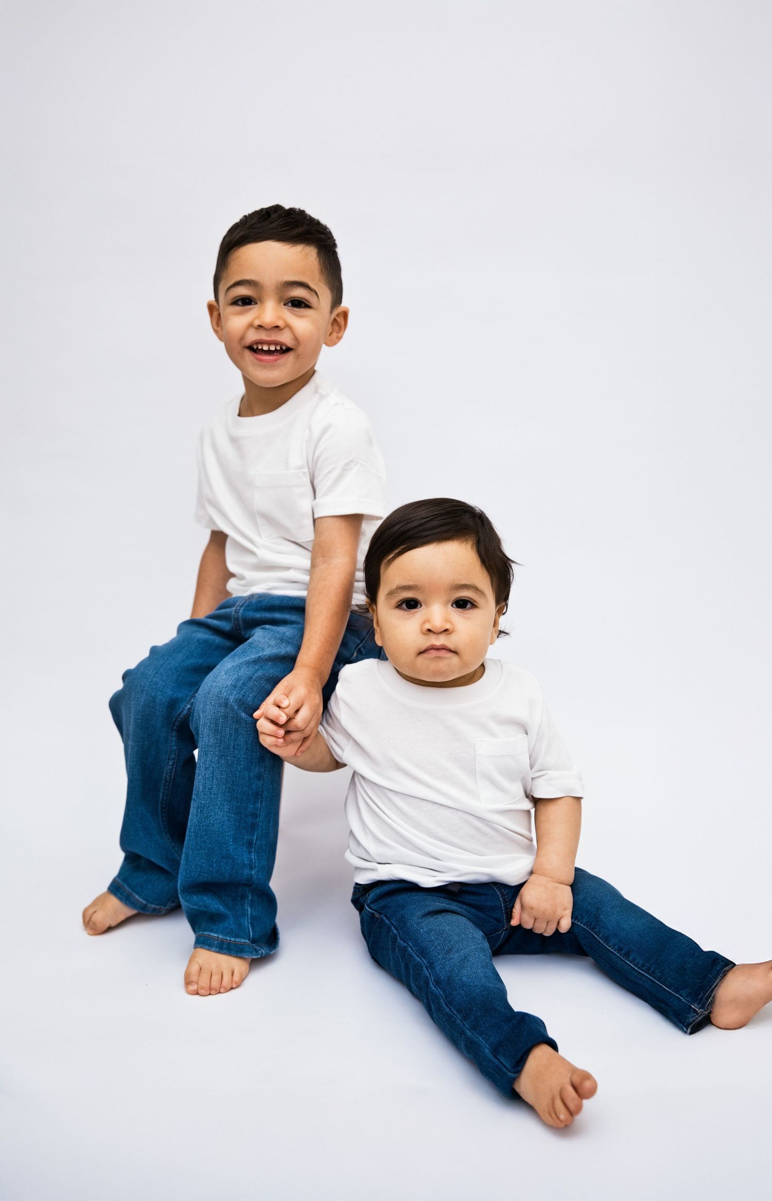 Two young brothers in matching white tees and jeans pose together, smiling, against a white studio background.