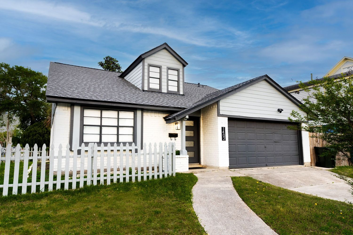 Charming white cottage-style home with gray roof, white picket fence, dormer window, and attached garage on a sunny day.