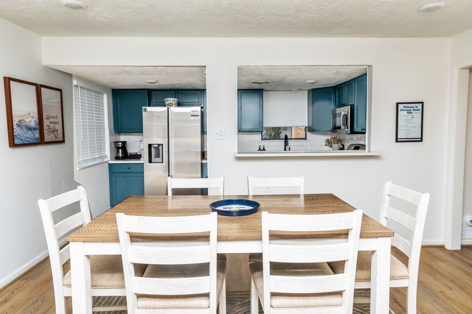 Bright dining area with wooden table, white chairs, and teal kitchen cabinets with stainless steel appliances visible.