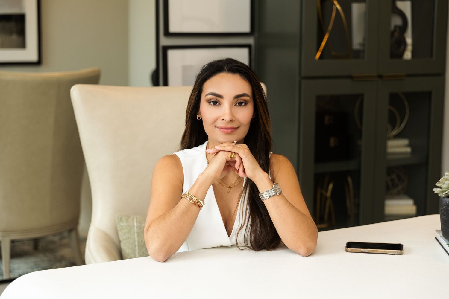 Professional woman in white sleeveless top sits at desk with hands clasped, smiling confidently in modern office.