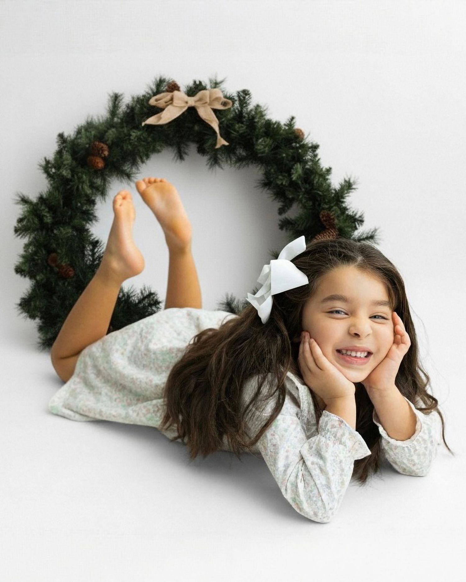 Child in white dress lies playfully near evergreen holiday wreath with gold bow and pinecone decorations. Katy TX christmas photos