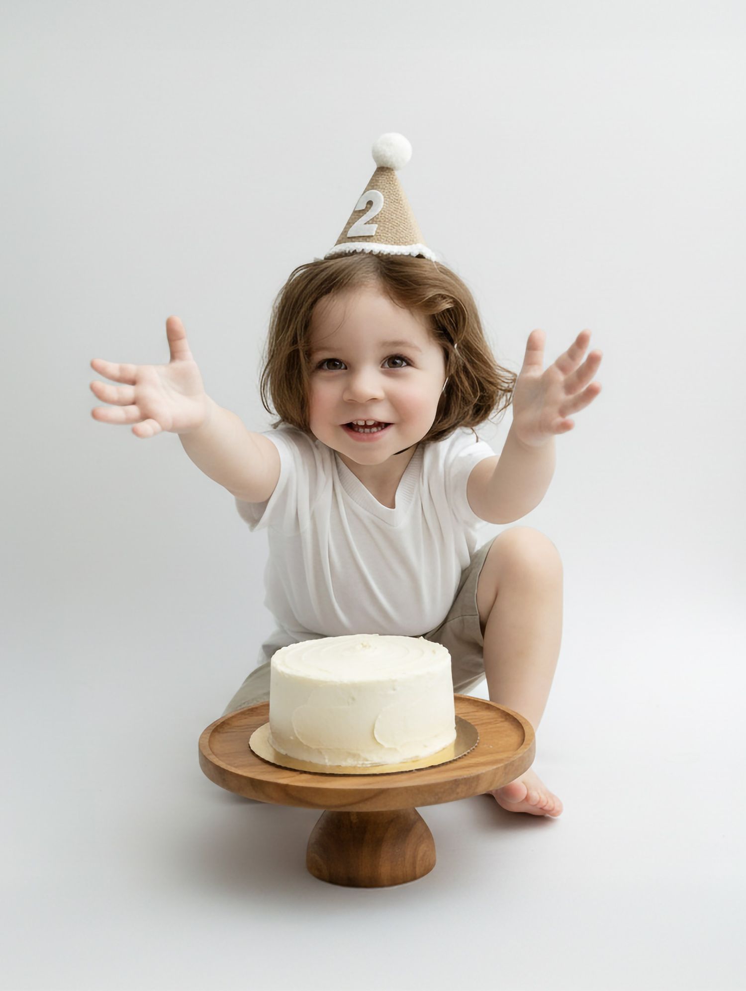 Child in white party dress gestures excitedly near white birthday cake on wooden stand.