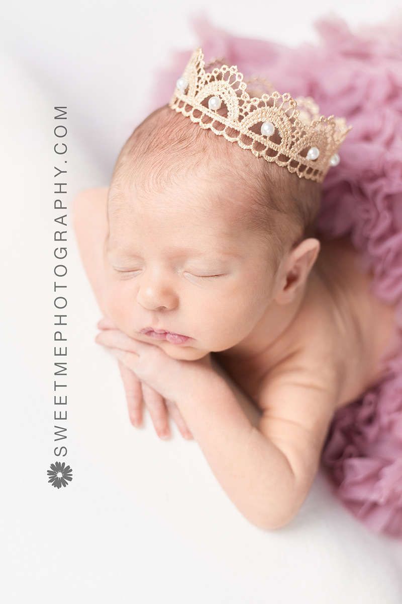 A sleeping newborn wearing a delicate gold crown tiara rests peacefully on soft pink fabric in a professional portrait.
