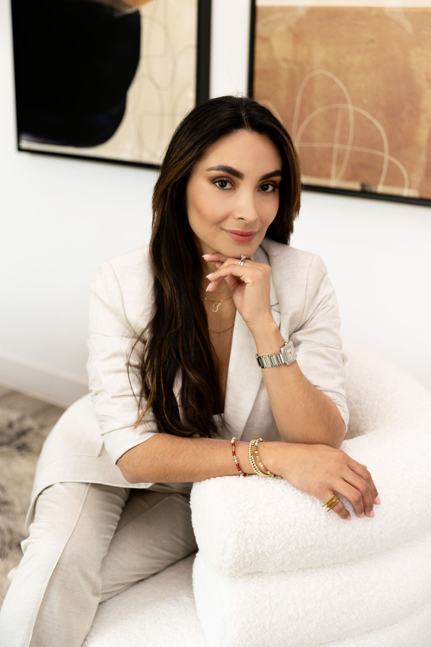 A stylish woman in a white blazer and pants sits elegantly on a white chair, smiling confidently in a modern interior.