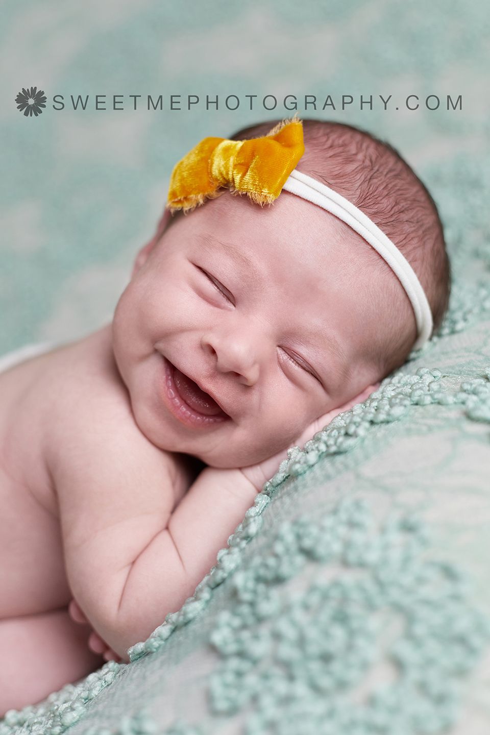 Newborn baby wearing yellow flower headband smiles while sleeping on mint green lace blanket.