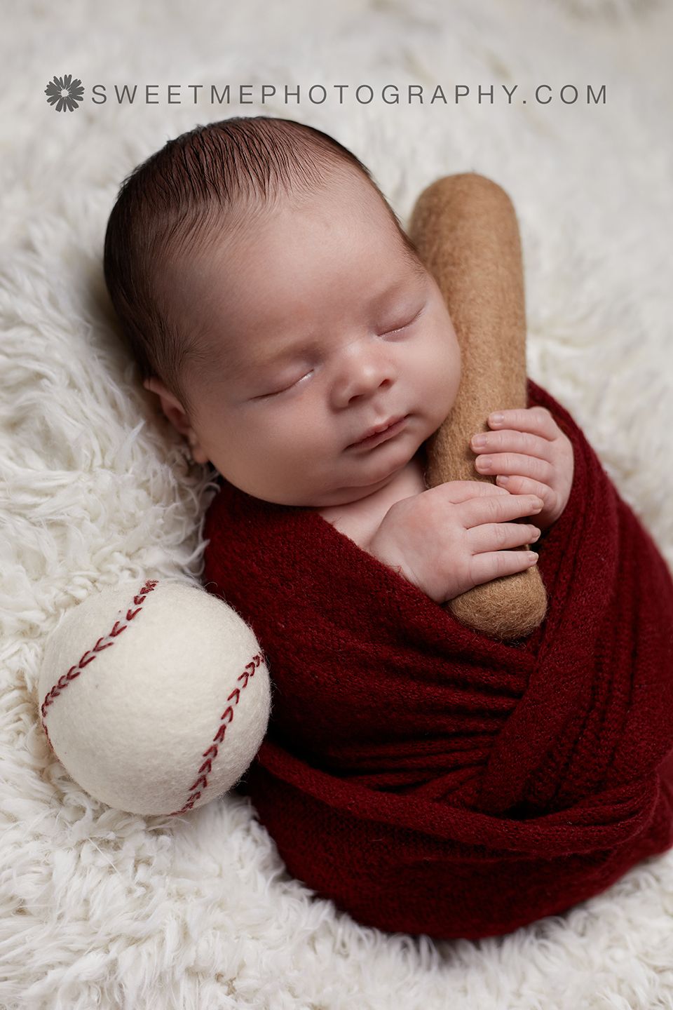 Sleeping newborn with baseball prop wrapped in burgundy fabric on white fluffy rug.