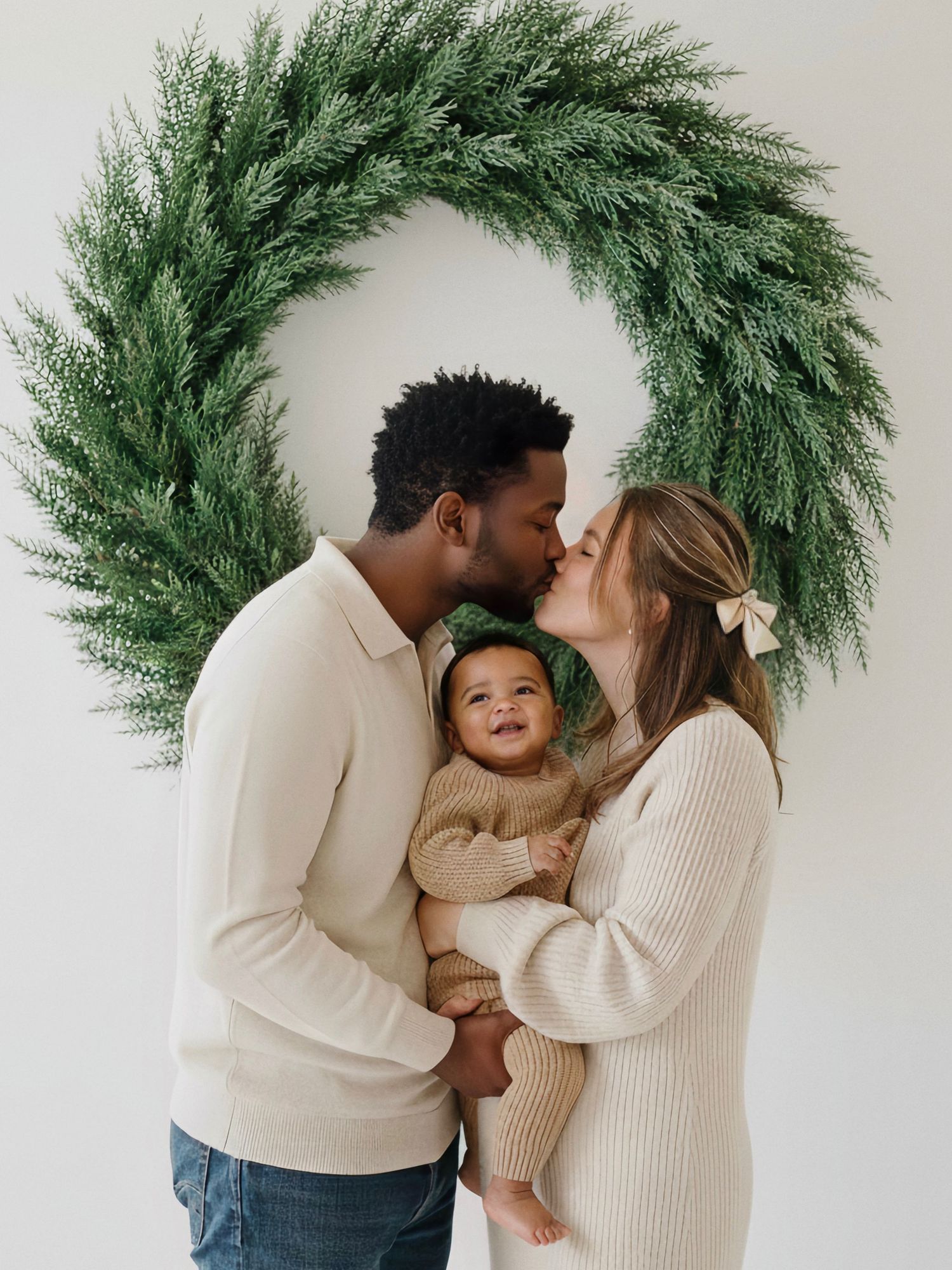 Family in matching cream sweaters share tender moments against white wall with evergreen wreath backdrop.