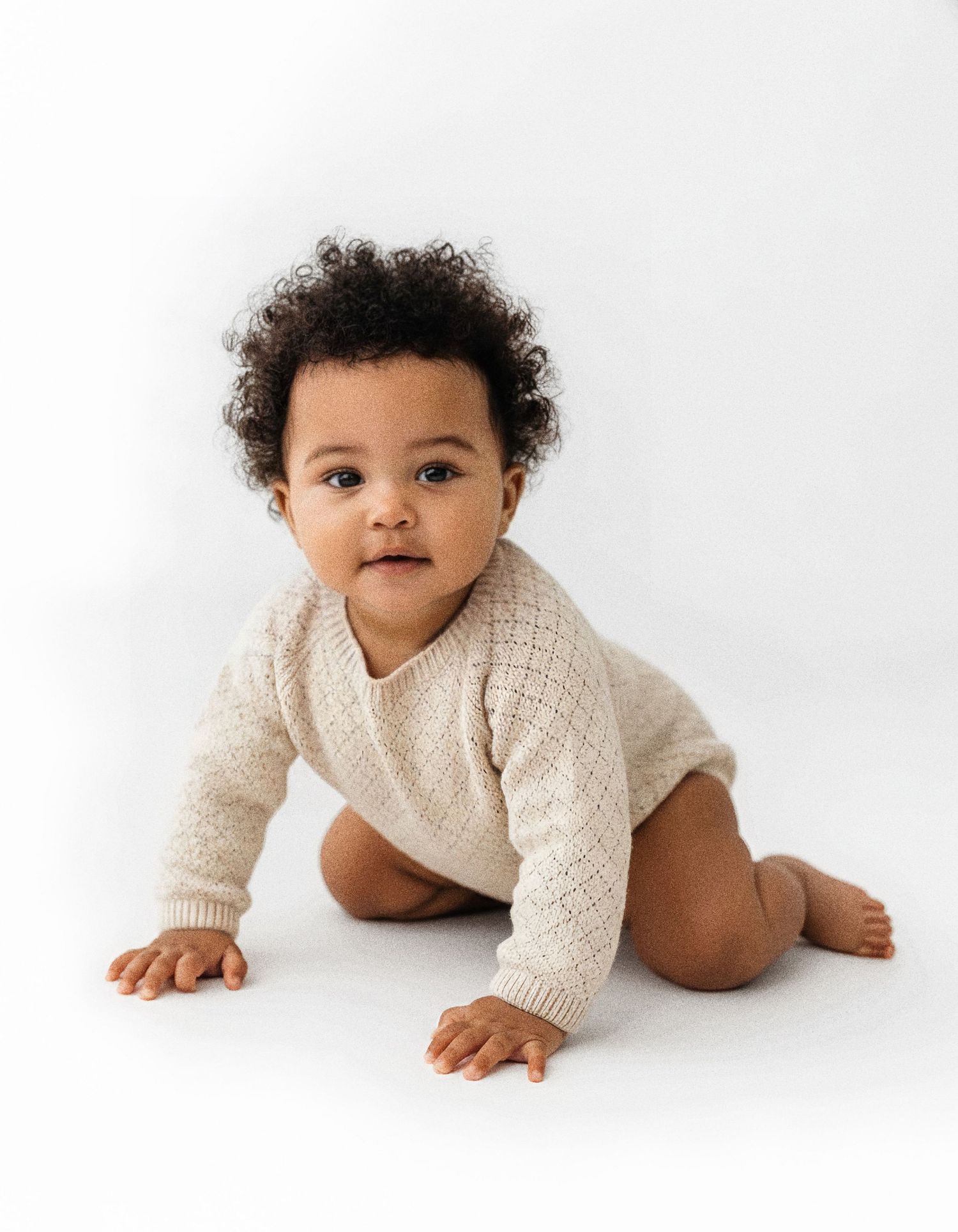 Baby wearing cream knit sweater crawls on white background while looking at camera.