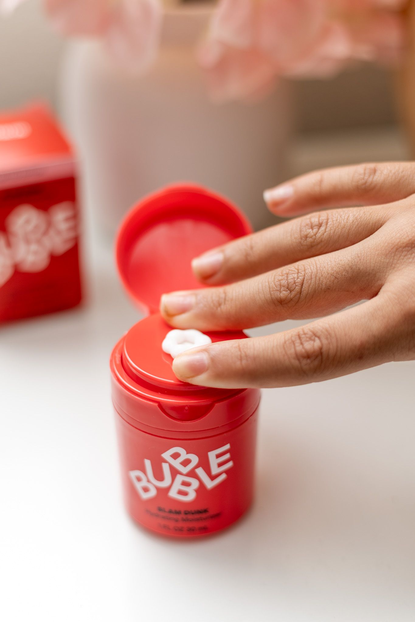 Hand dipping finger into Bubble Skincare red Slam Dunk moisturizer jar on white surface with pink flowers.