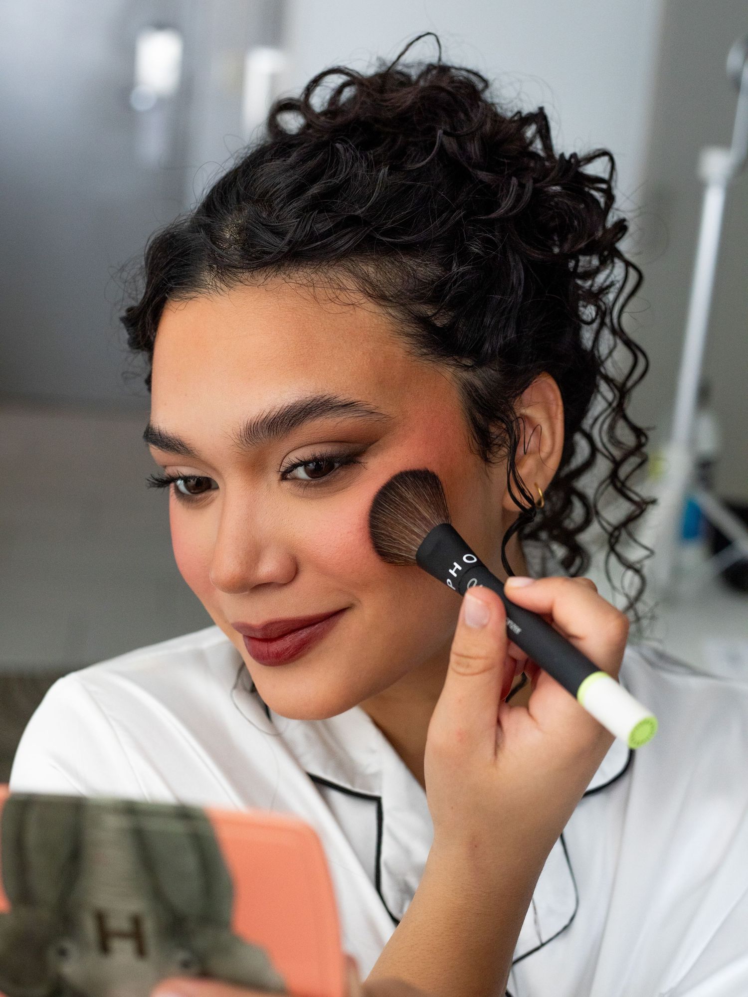 A makeup artist applies blush to a model's cheeks using a fluffy brush in a bright studio setting.