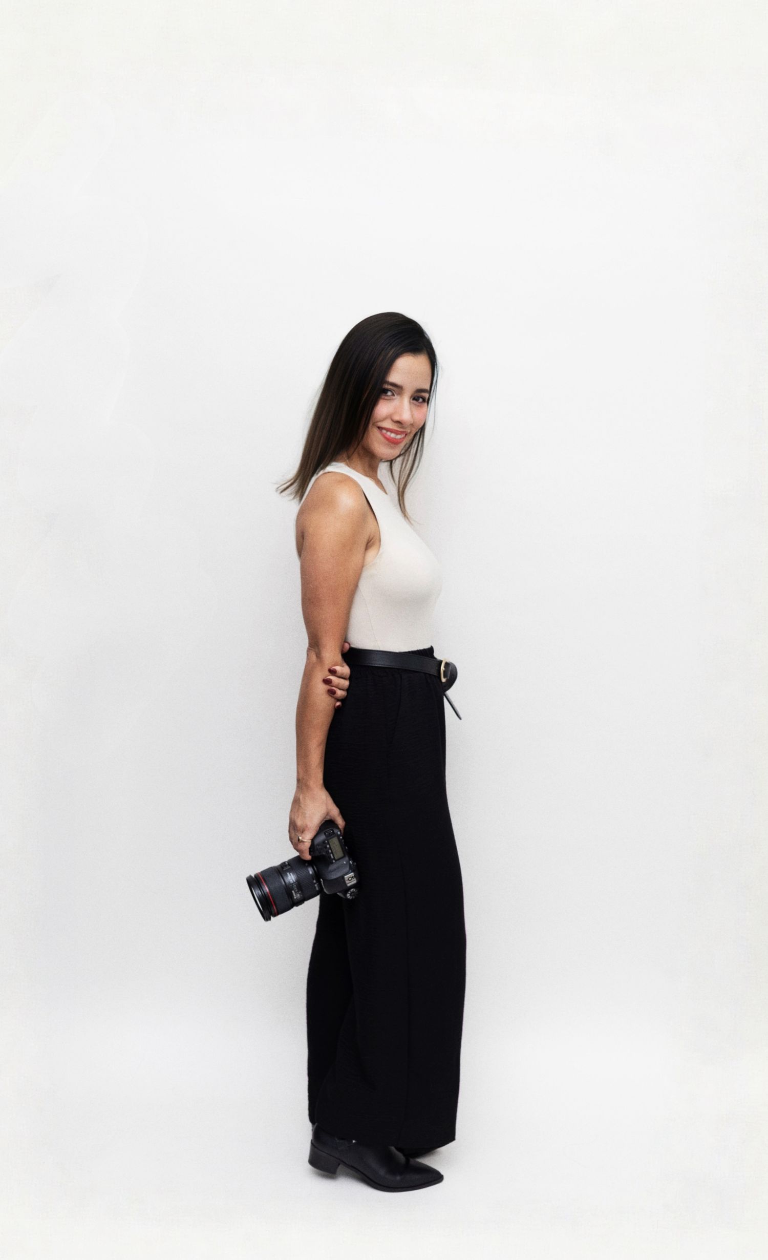 Professional photographer in a black dress poses with camera equipment against a white studio backdrop.