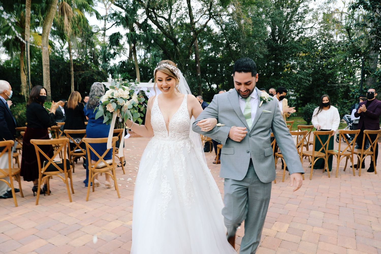 A wedding couple dances together at an outdoor reception surrounded by palm trees and wooden chairs.
