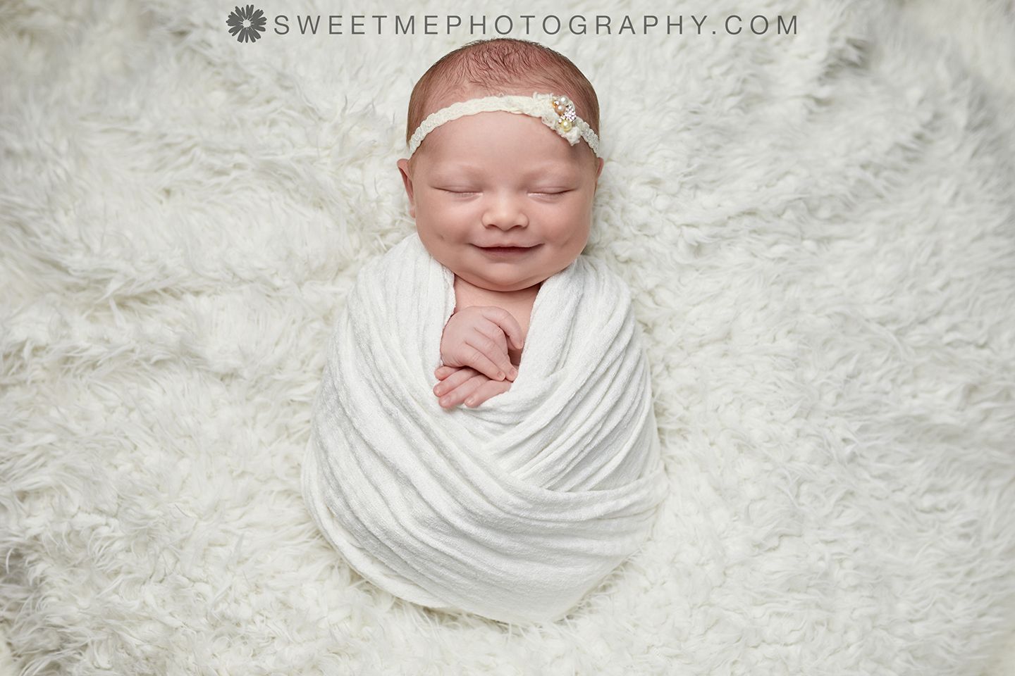 Newborn baby wrapped in soft white fabric lying on textured white blanket wearing delicate headband.