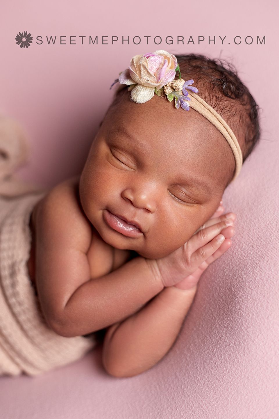 Peaceful newborn sleeping on pink backdrop wearing delicate floral headband in professional portrait.