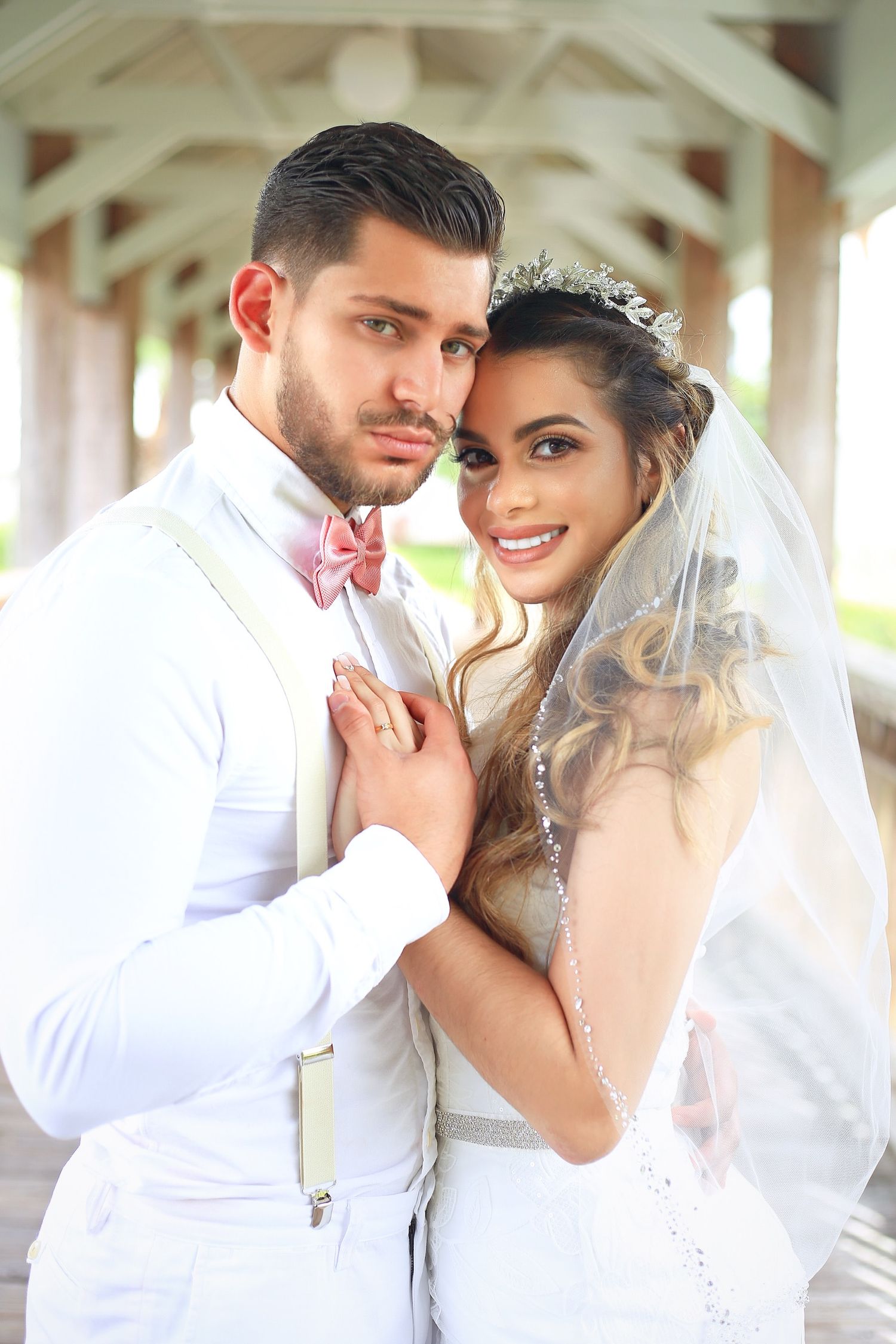 Wedding portrait of couple in all white attire with bride wearing veil and groom in bow tie under covered walkway.