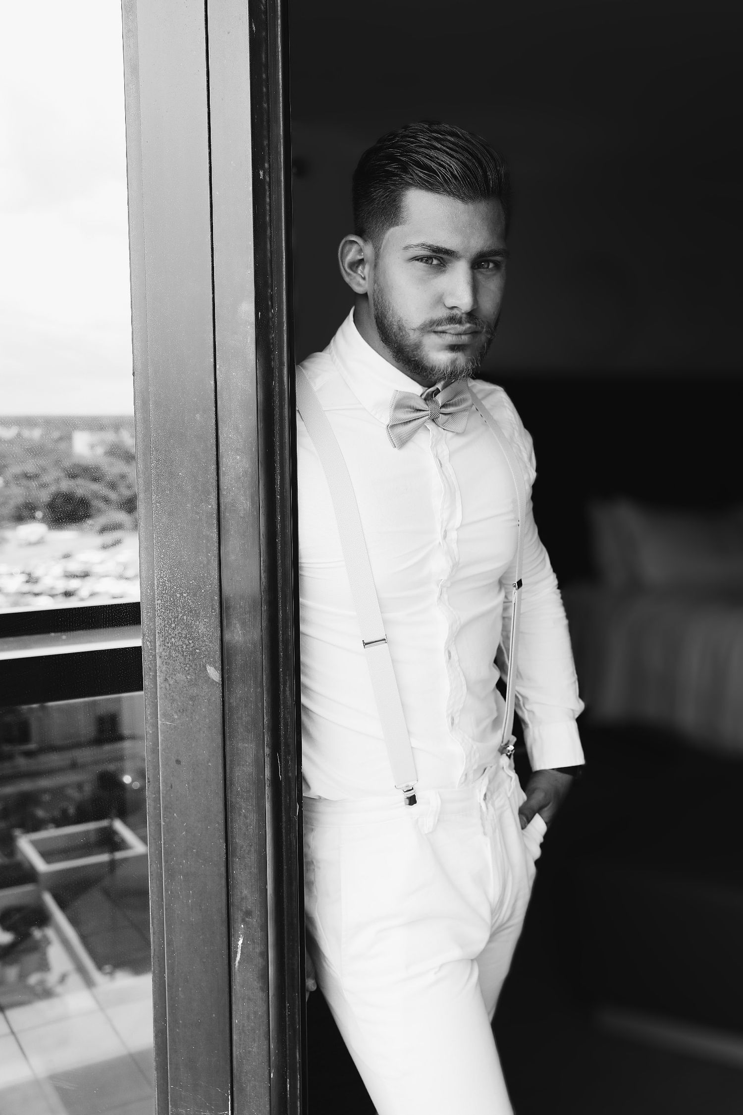 Black and white portraits of a groom in a white dress shirt and bow tie looking out a window.