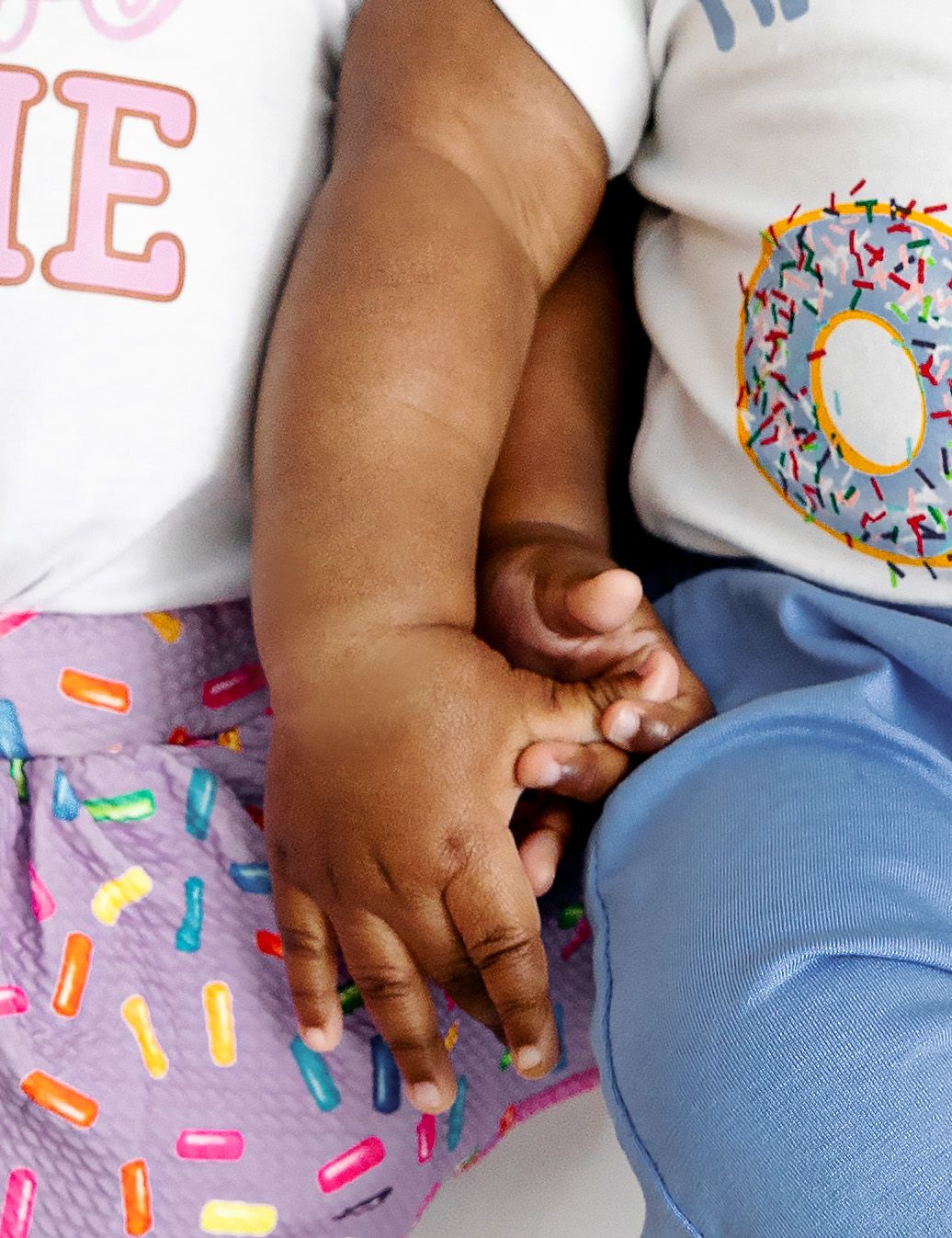 Two babies holding hands wearing donut-themed outfits with colorful sprinkle print clothing.