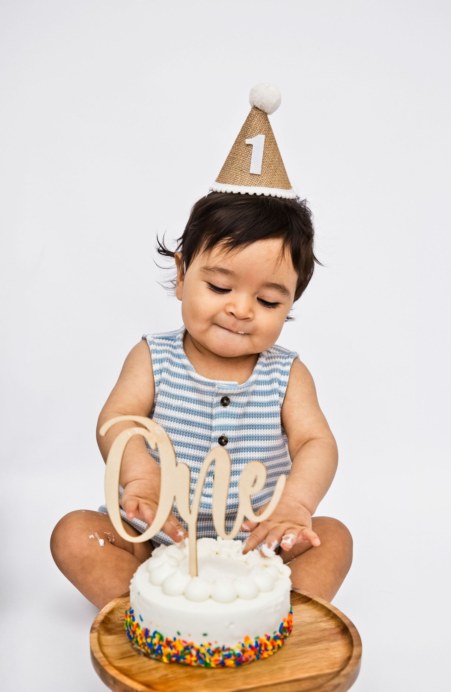 Baby's first birthday smash cake photo with a number one party hat, blue striped outfit, and colorful sprinkle cake.