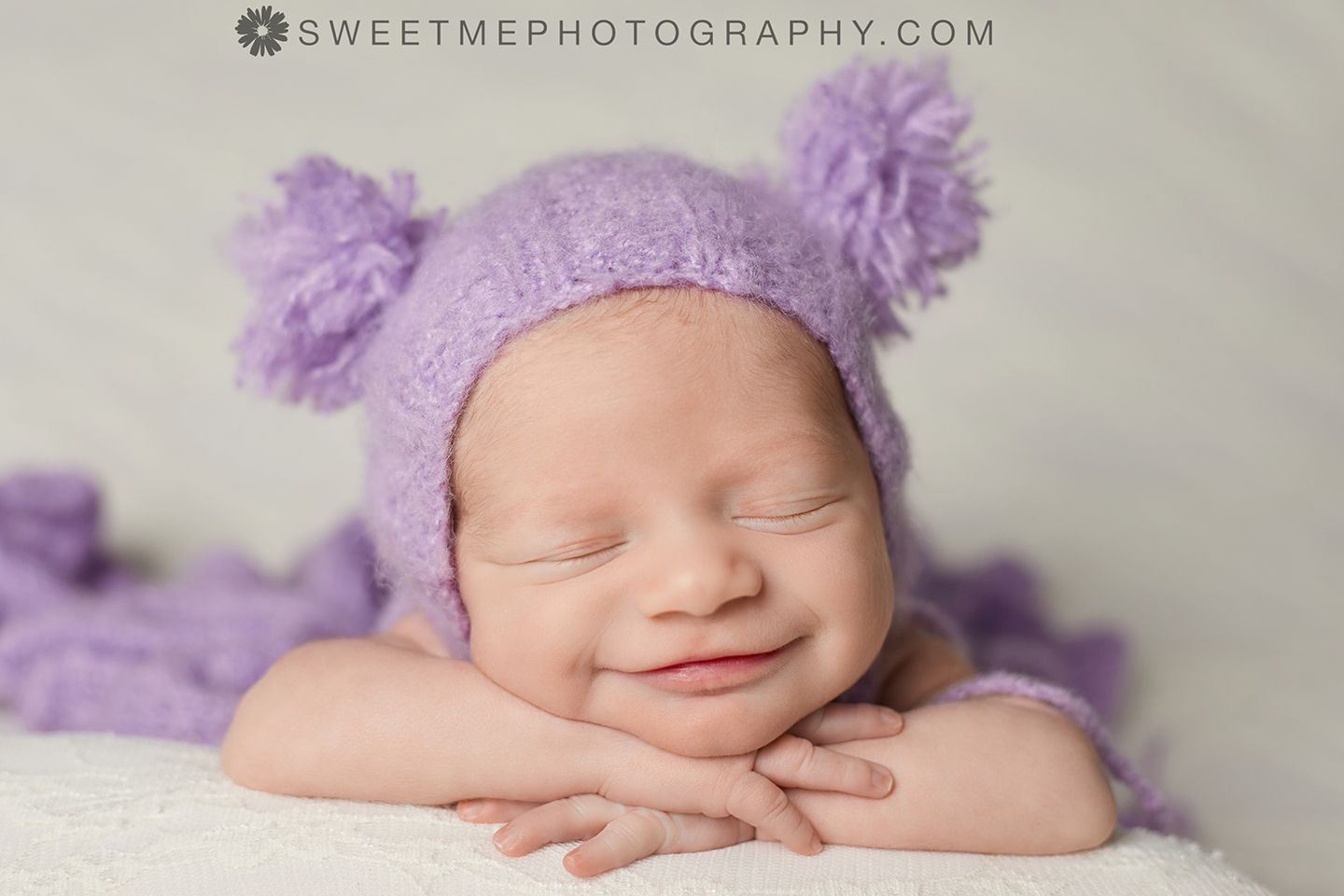 Sleeping newborn in purple knit hat and wrap on soft gray blanket in professional studio photography.