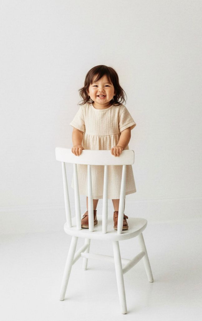 A small child in a cream-colored dress stands joyfully on a white wooden chair against a light background.