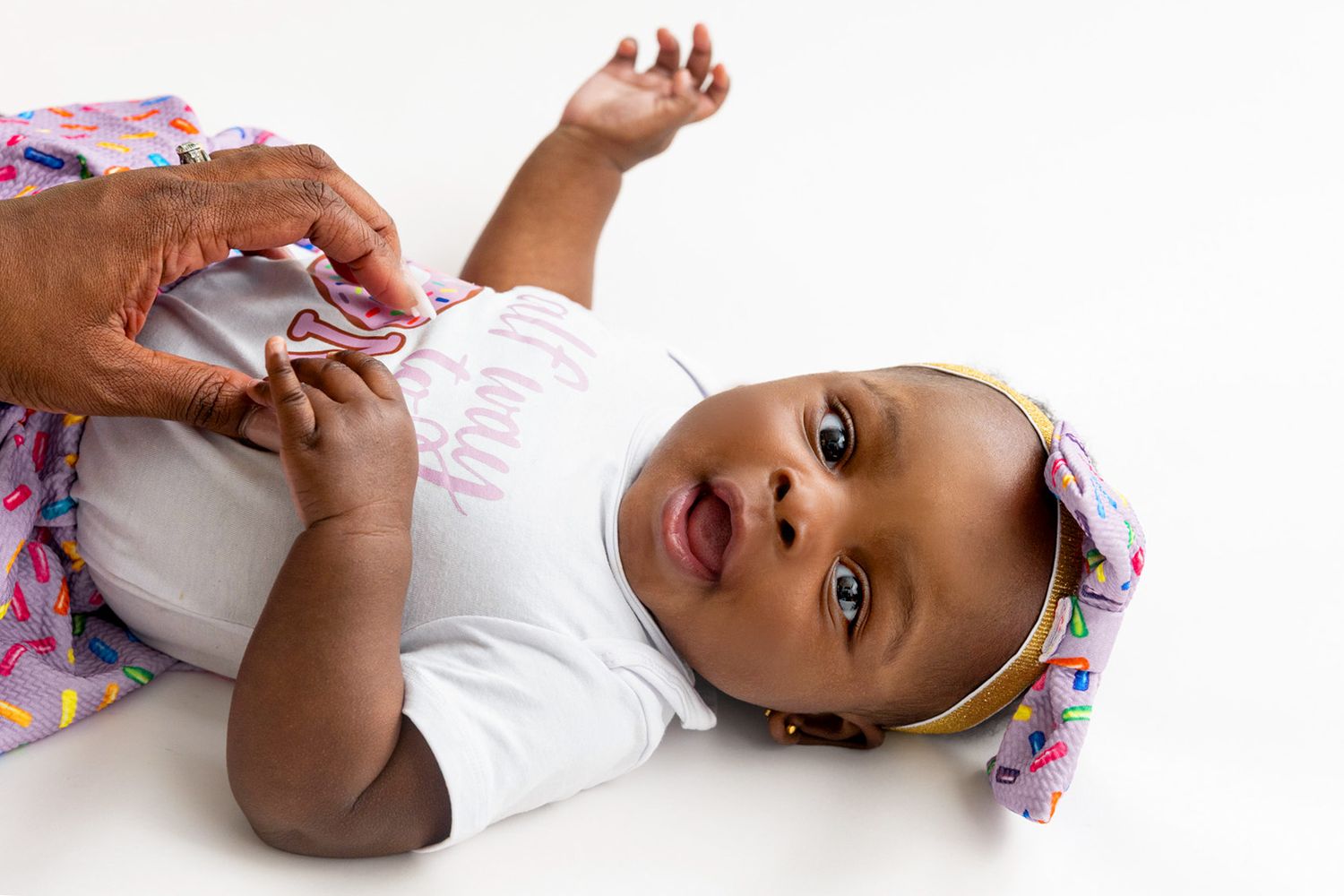Adorable Black baby girl lying down wearing a white onesie and gold headband, smiling while an adult adjusts her outfit.