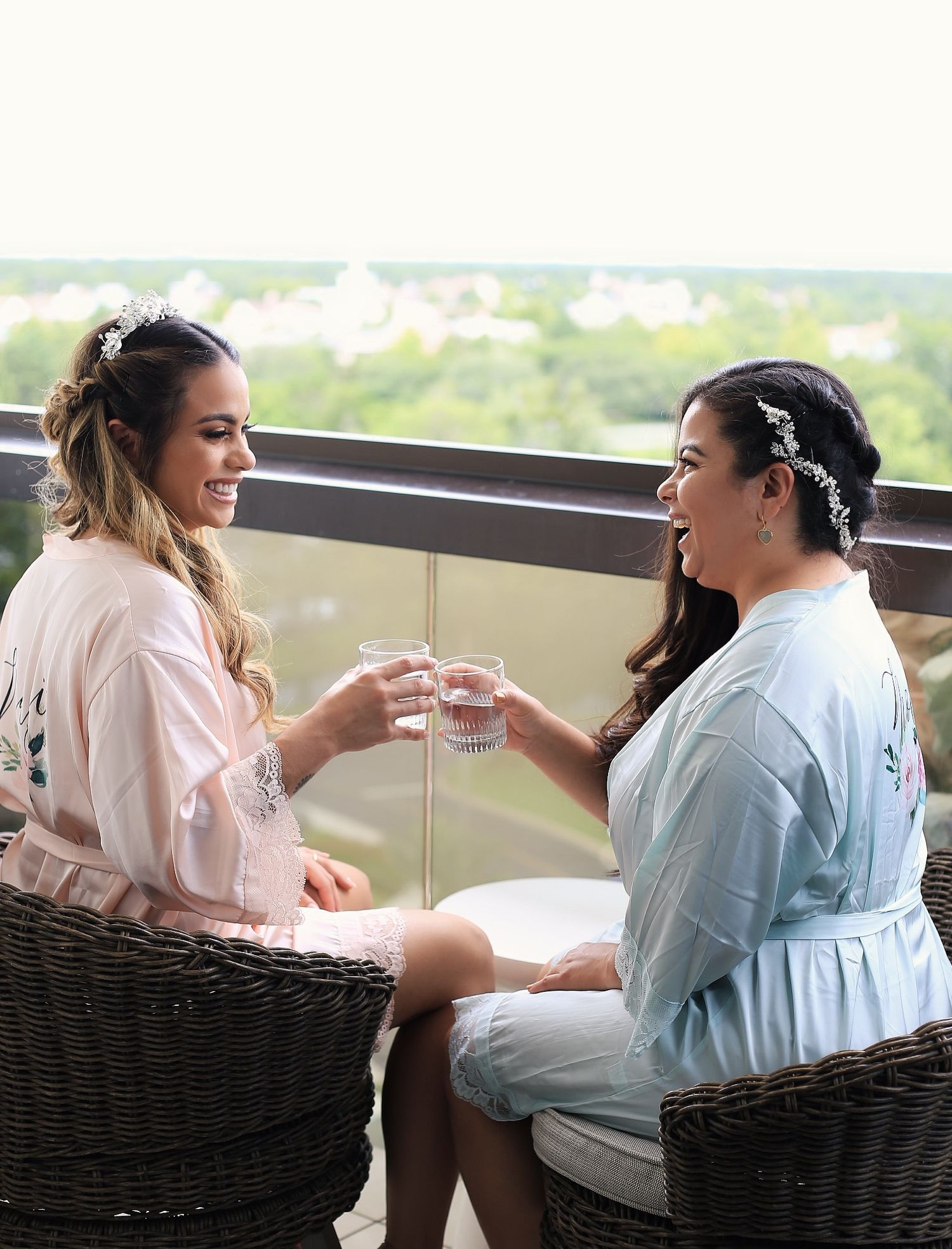 Two women in robes toast with glasses while sitting in wicker chairs on a balcony overlooking a scenic view.