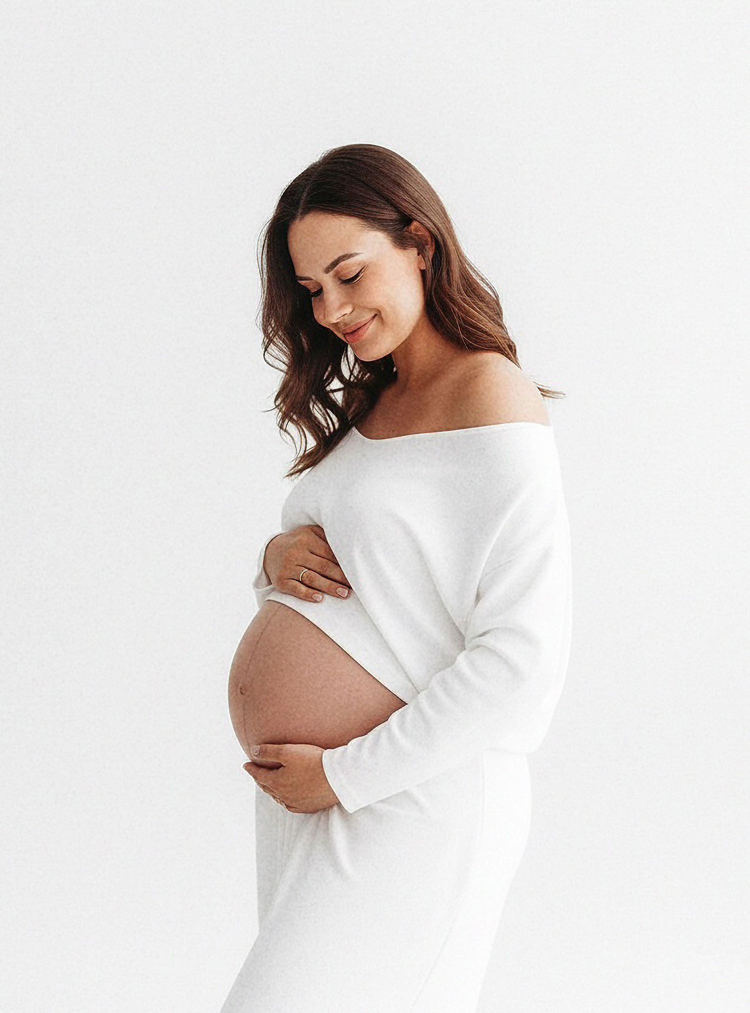 Pregnant woman in white sweater poses against white background in three similar maternity photos.