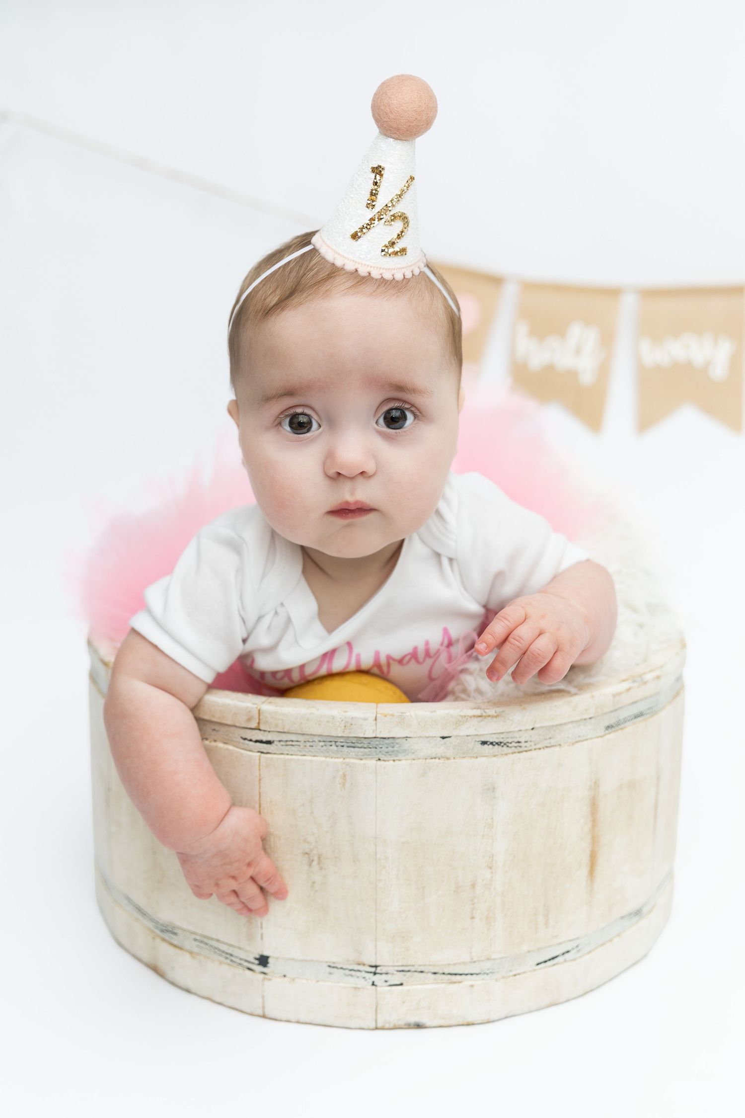 Baby girl in white onesie and half birthday party hat sits in wooden barrel with pink tutu.