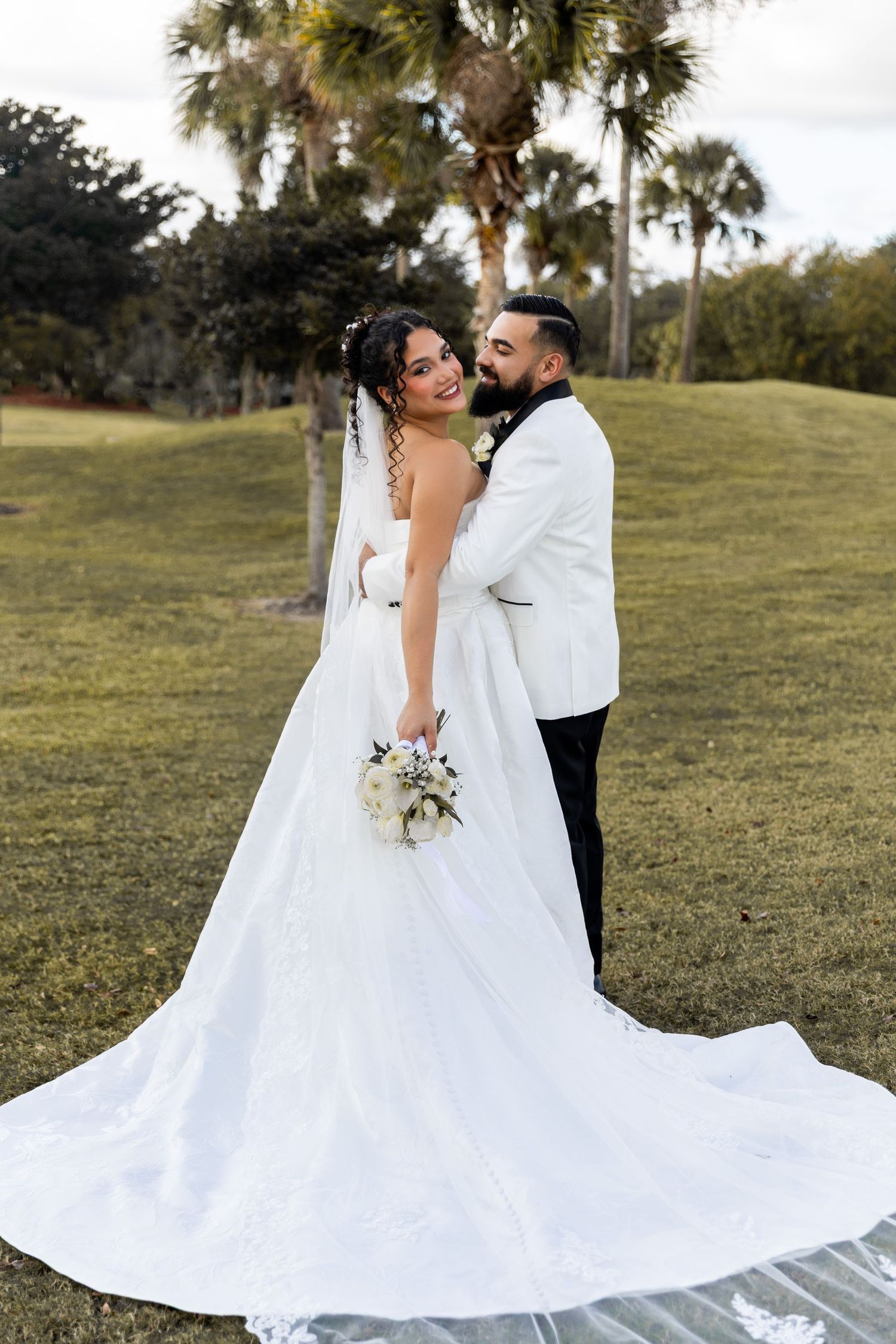 A romantic wedding couple embraces on a grassy hillside with palm trees in the background.