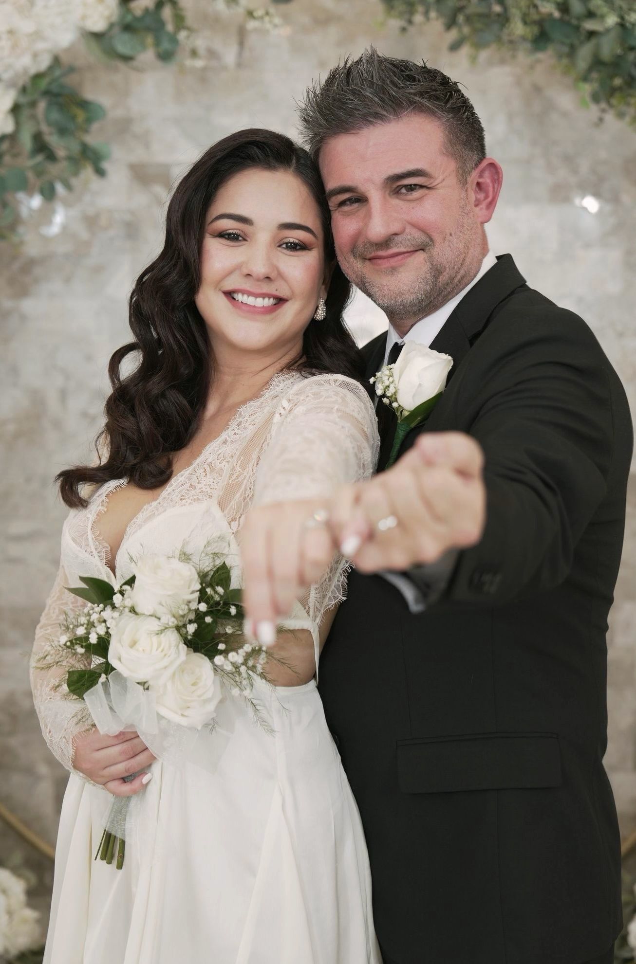 Joyful wedding couple poses together against stone wall with white floral bouquet.