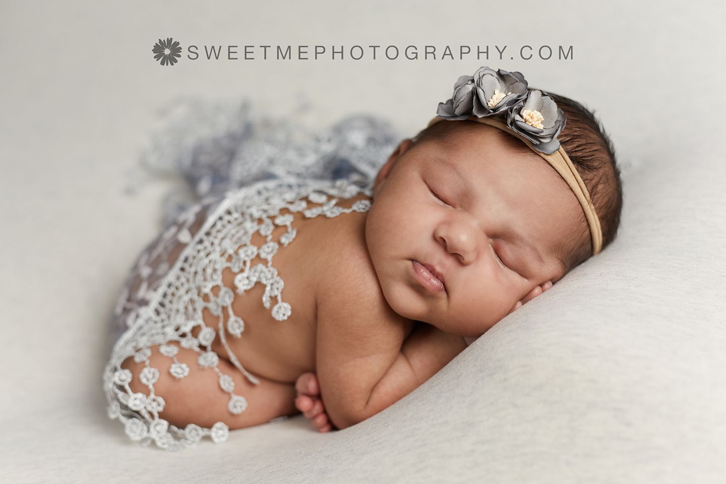 A sleeping newborn baby wearing a delicate lace headband rests on soft white fabric.