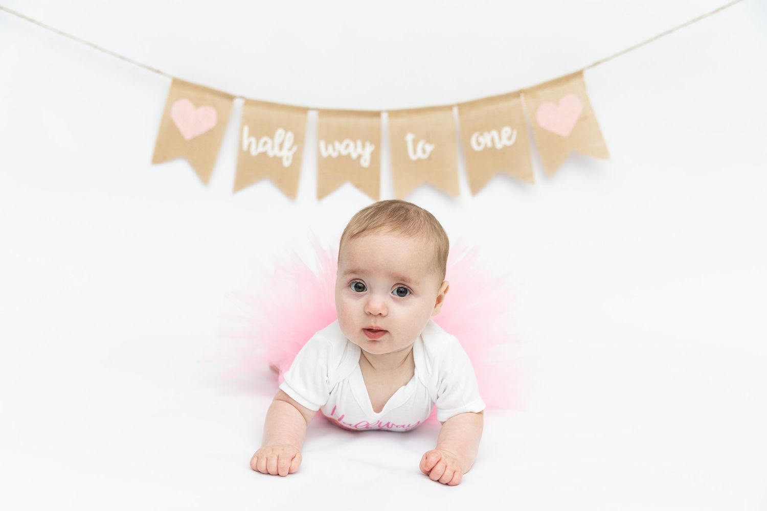 Baby girl in pink tutu lies on white floor under a burlap “half way to one” banner with pink hearts.