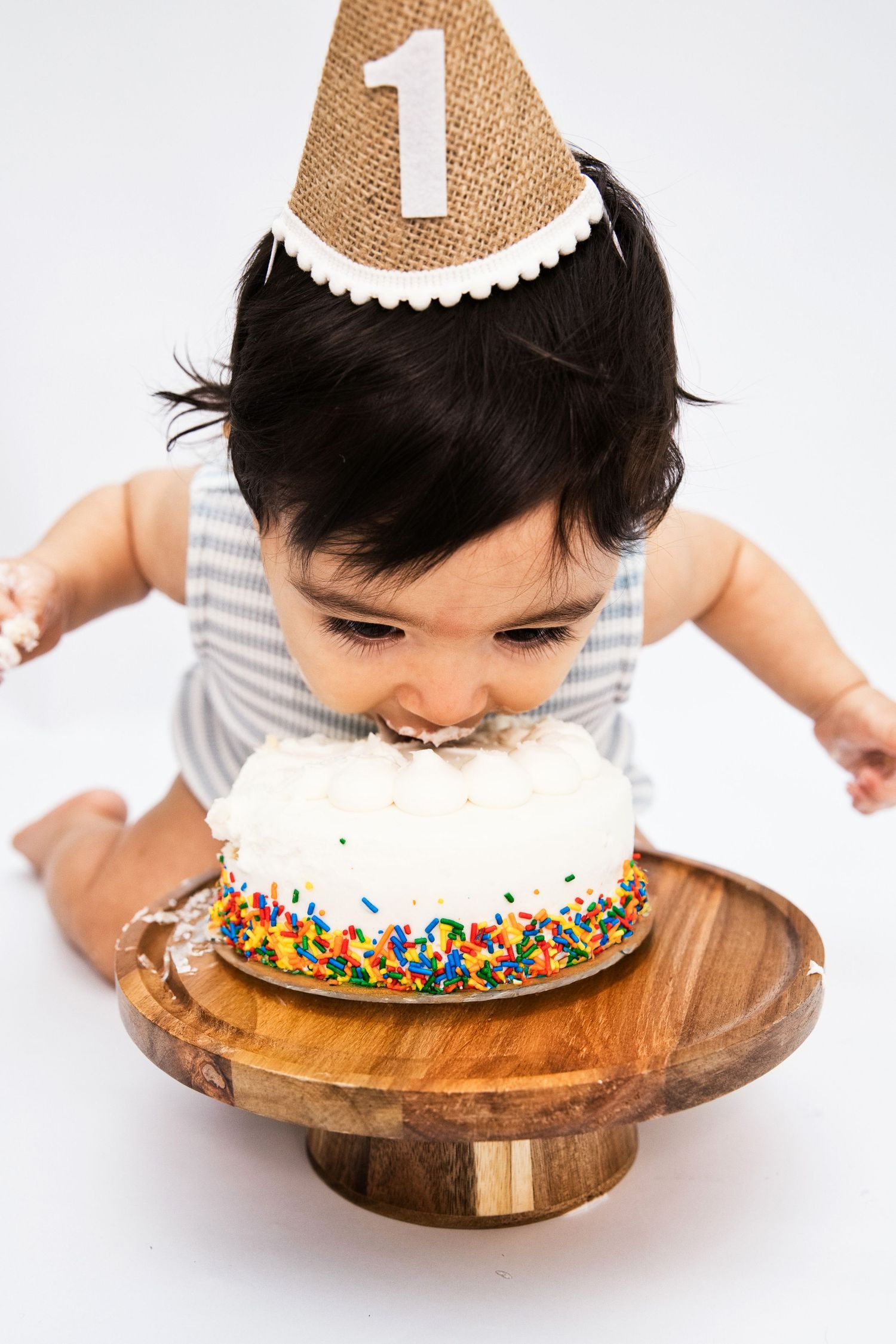 A baby wearing a number one party hat smashes their face into a white sprinkle-covered first birthday cake on a wooden stand.