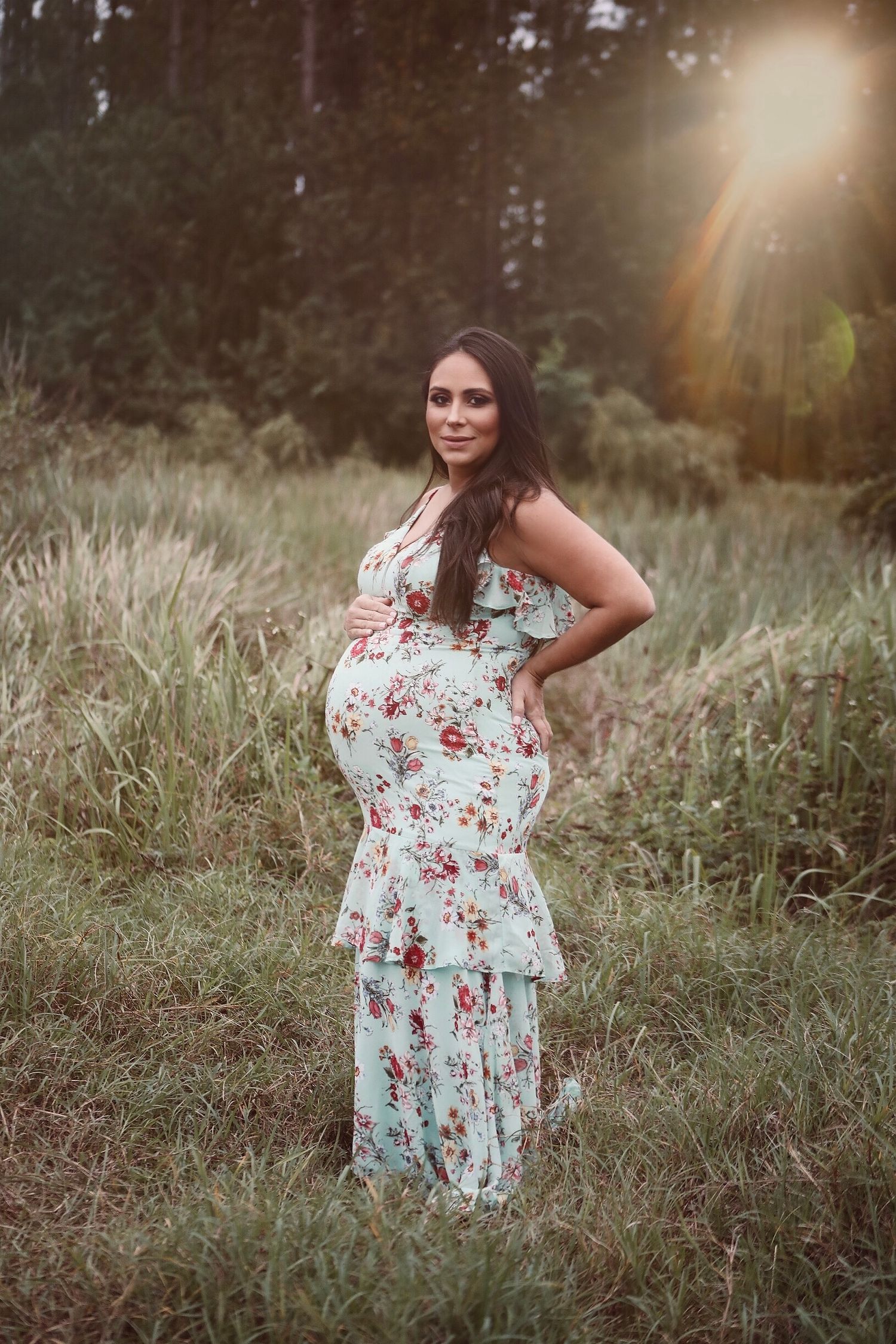 A floral maternity dress photoshoot in a sunlit meadow captures a glowing moment during golden hour.