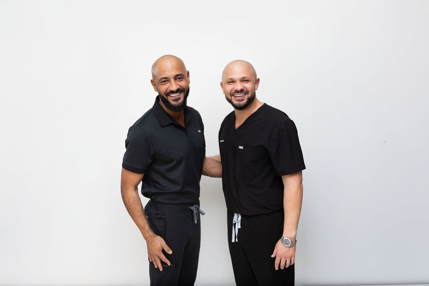Two smiling men in matching black scrubs pose together against a white background in a professional healthcare setting.