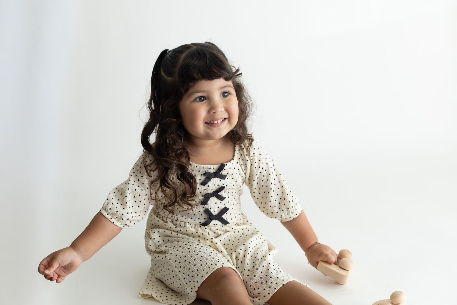 Child in polka dot dress with black bow sits and plays on white backdrop.