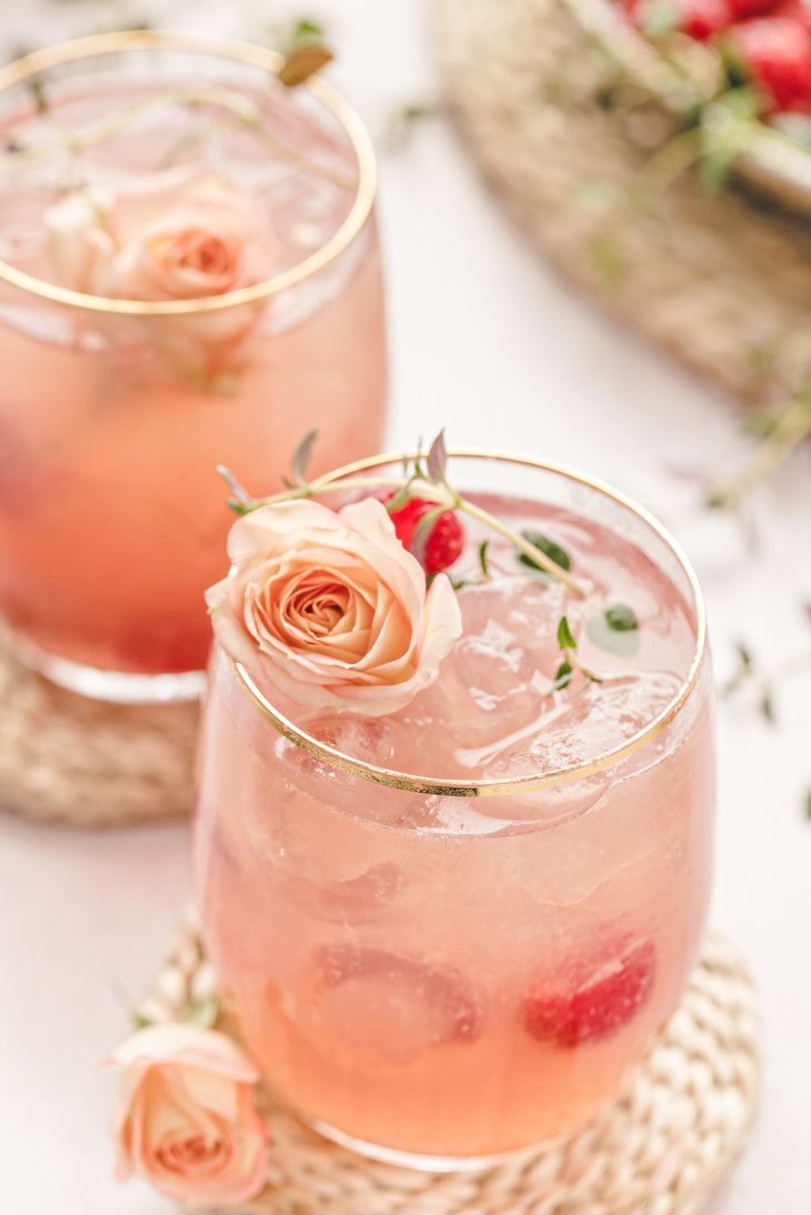 Two pink cocktails garnished with peach roses and raspberries on woven coasters against a light background.